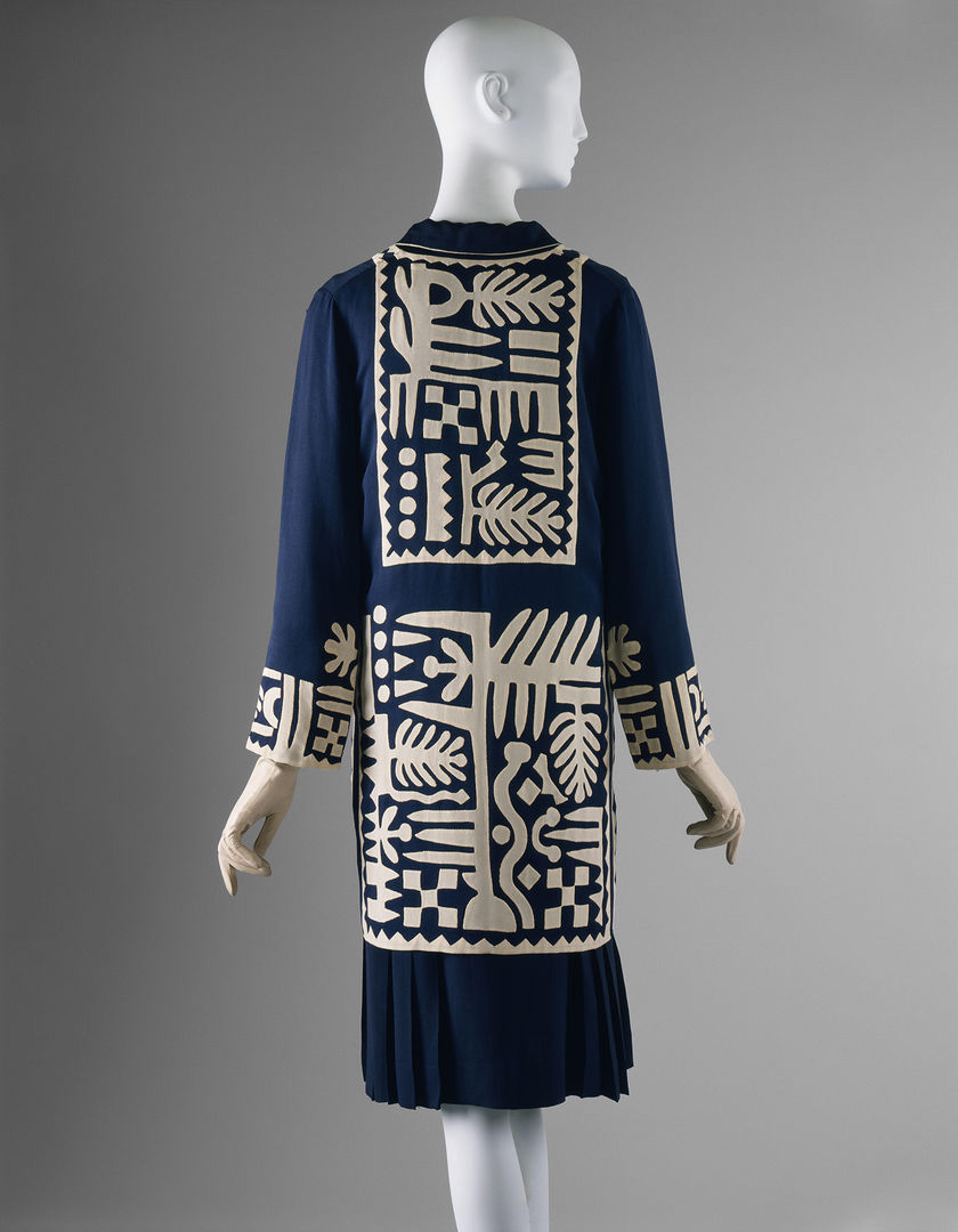 The back of a navy blue dress with beautiful white patterning on the back and lower part of the dress and around the wrists. The dress is against a grey background and fitted onto a mannequin with a head and hands.