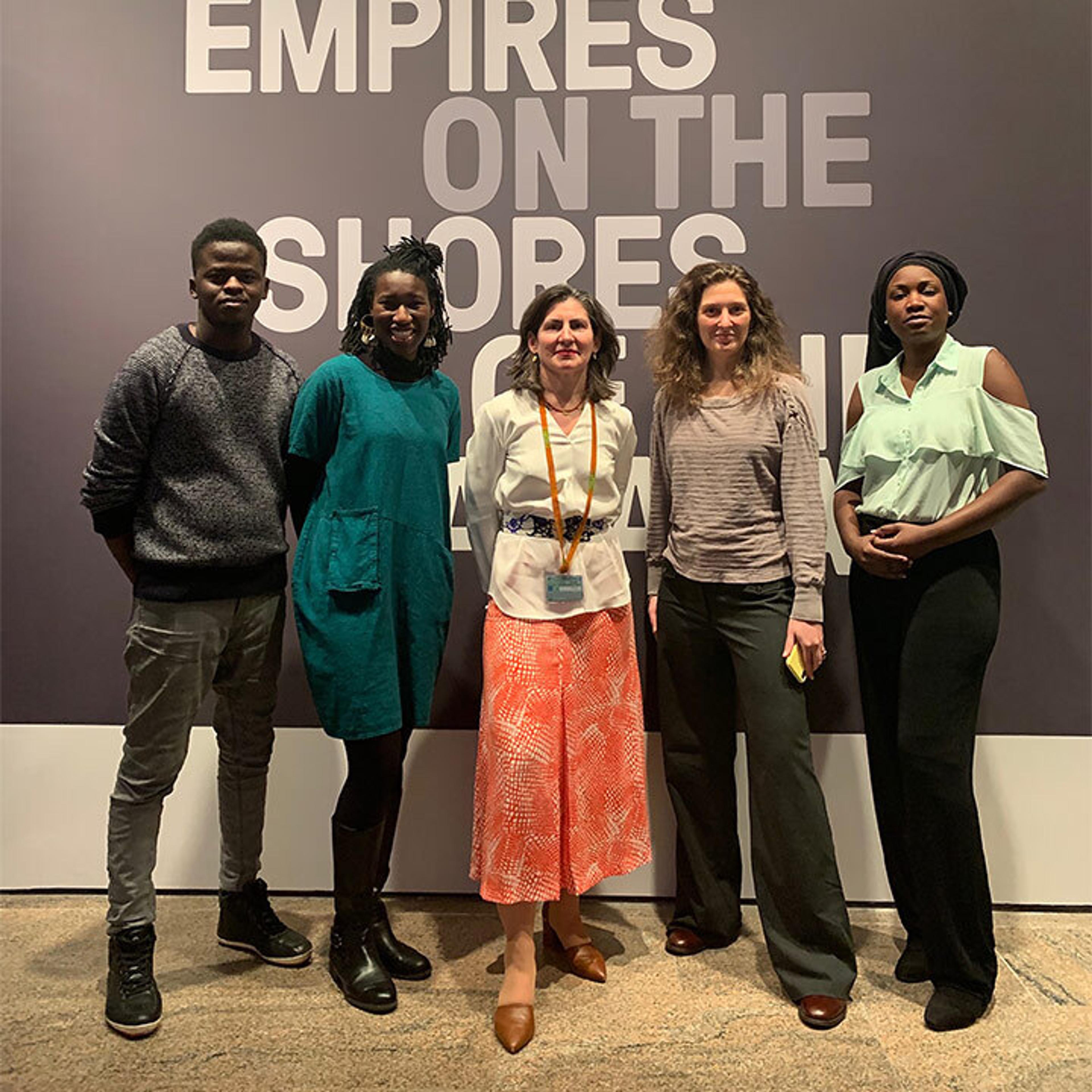 Five people smile and stand in front of an ivory wall with white text that reads: "On the Shores"