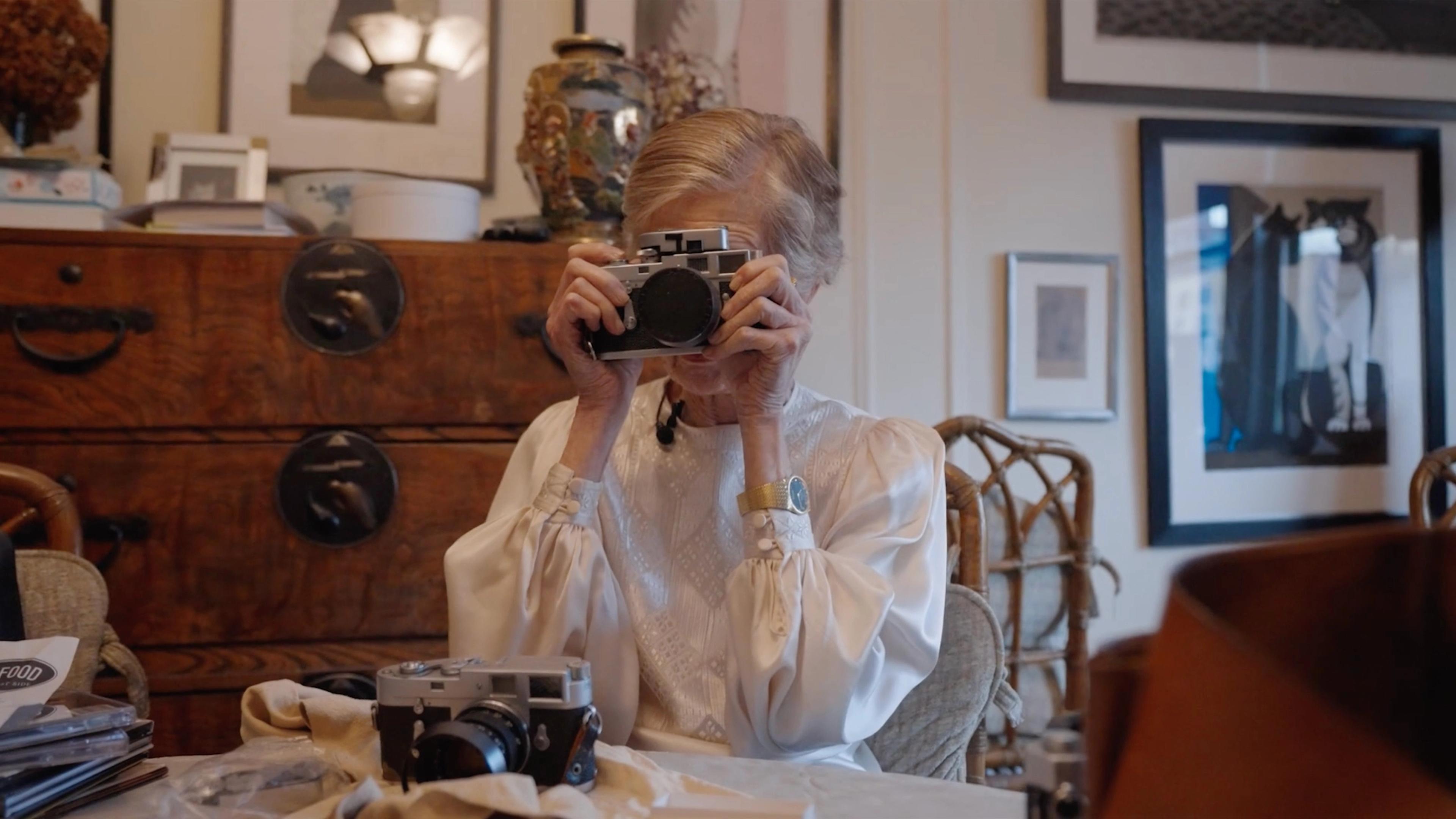 A woman seated at a table holds a camera in front of her face.