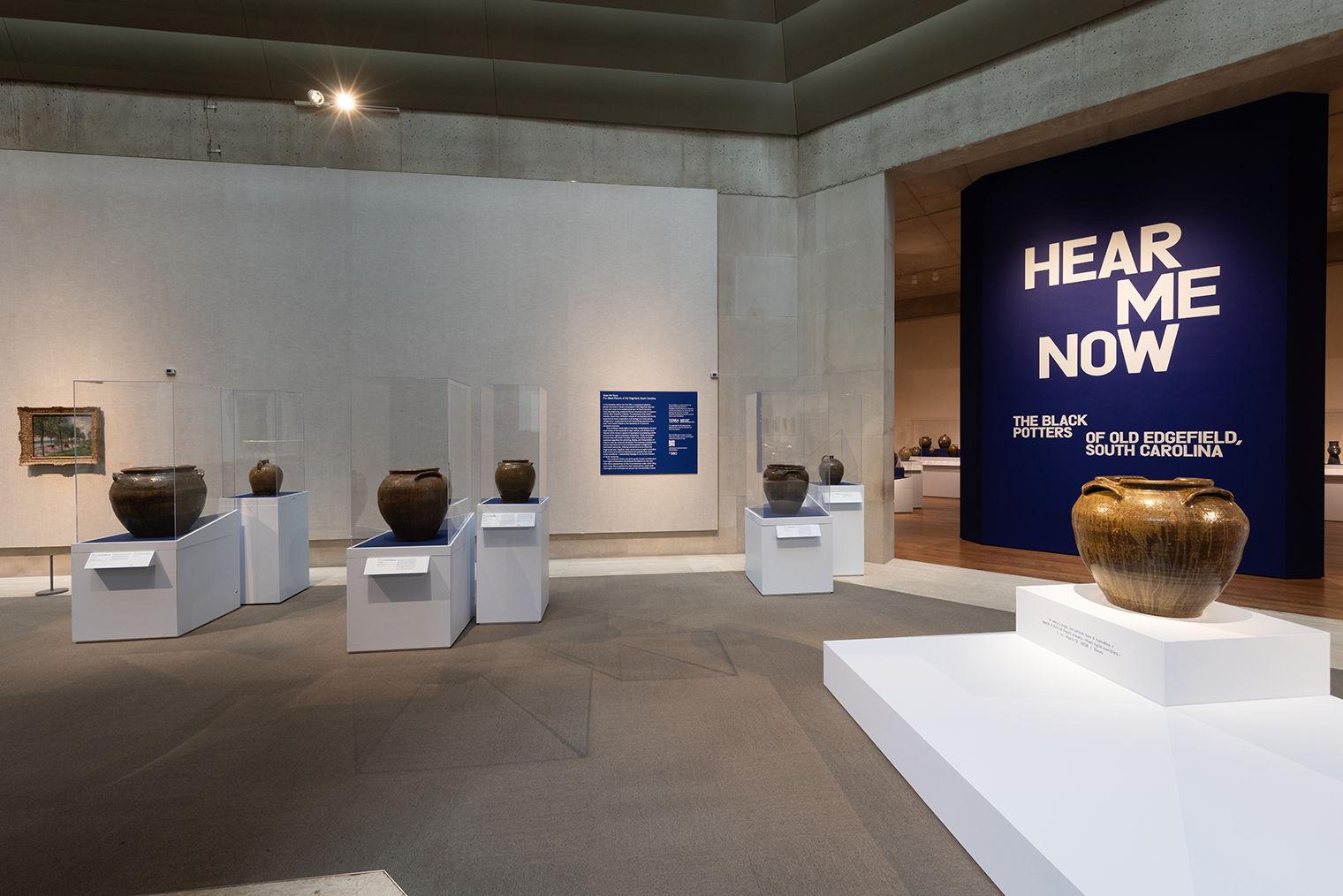 Shot of the galleries for the exhibition Hear Me Now: The Black Potters of Old Edgefield, South Carolina with several of the jars from the show on white plinths.