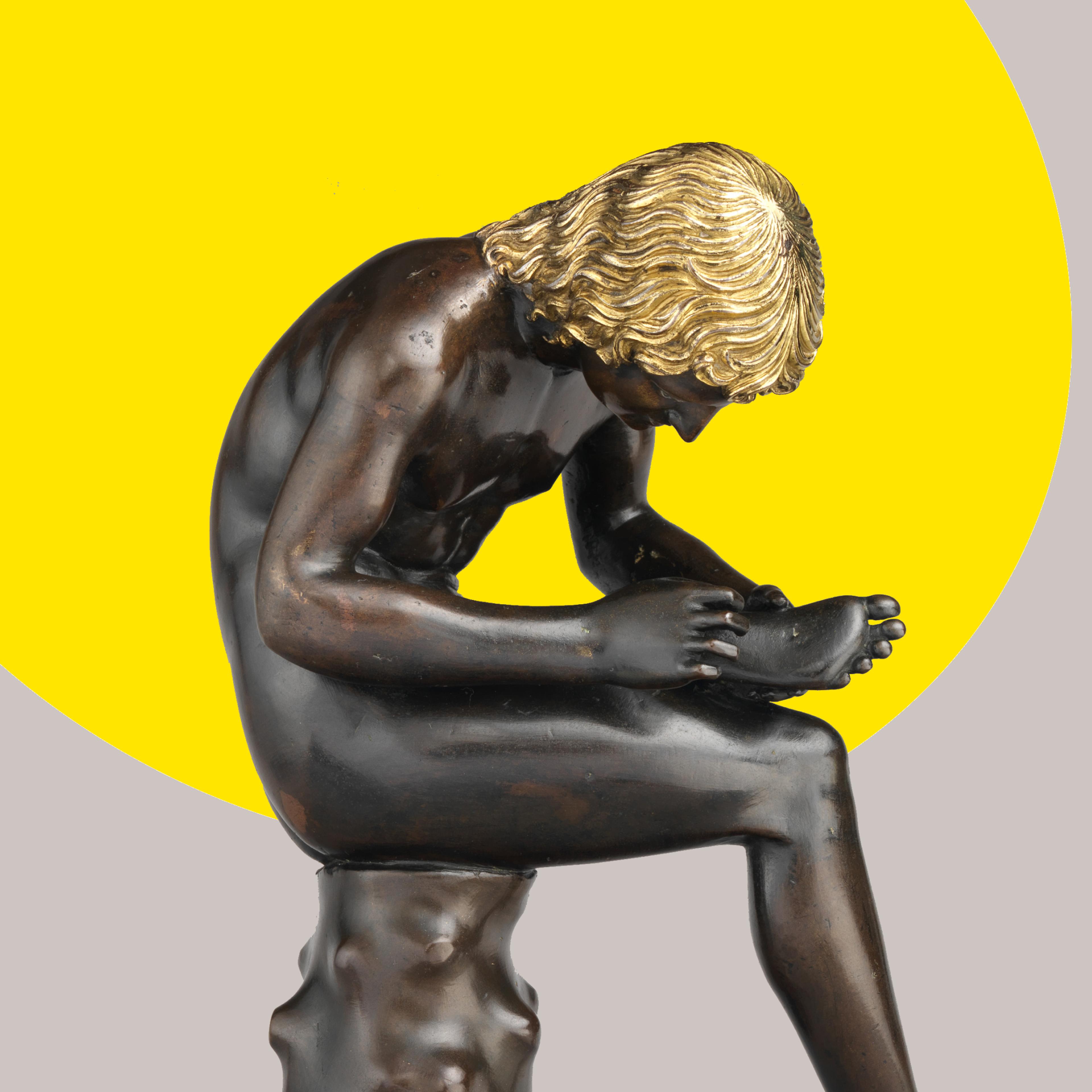 A small bronze sculpture of a seated boy pulling a thorn from the bottom of his foot set against a gray-scale studio backdrop. A bright yellow ovular spotlight shape appears from behind the figure.