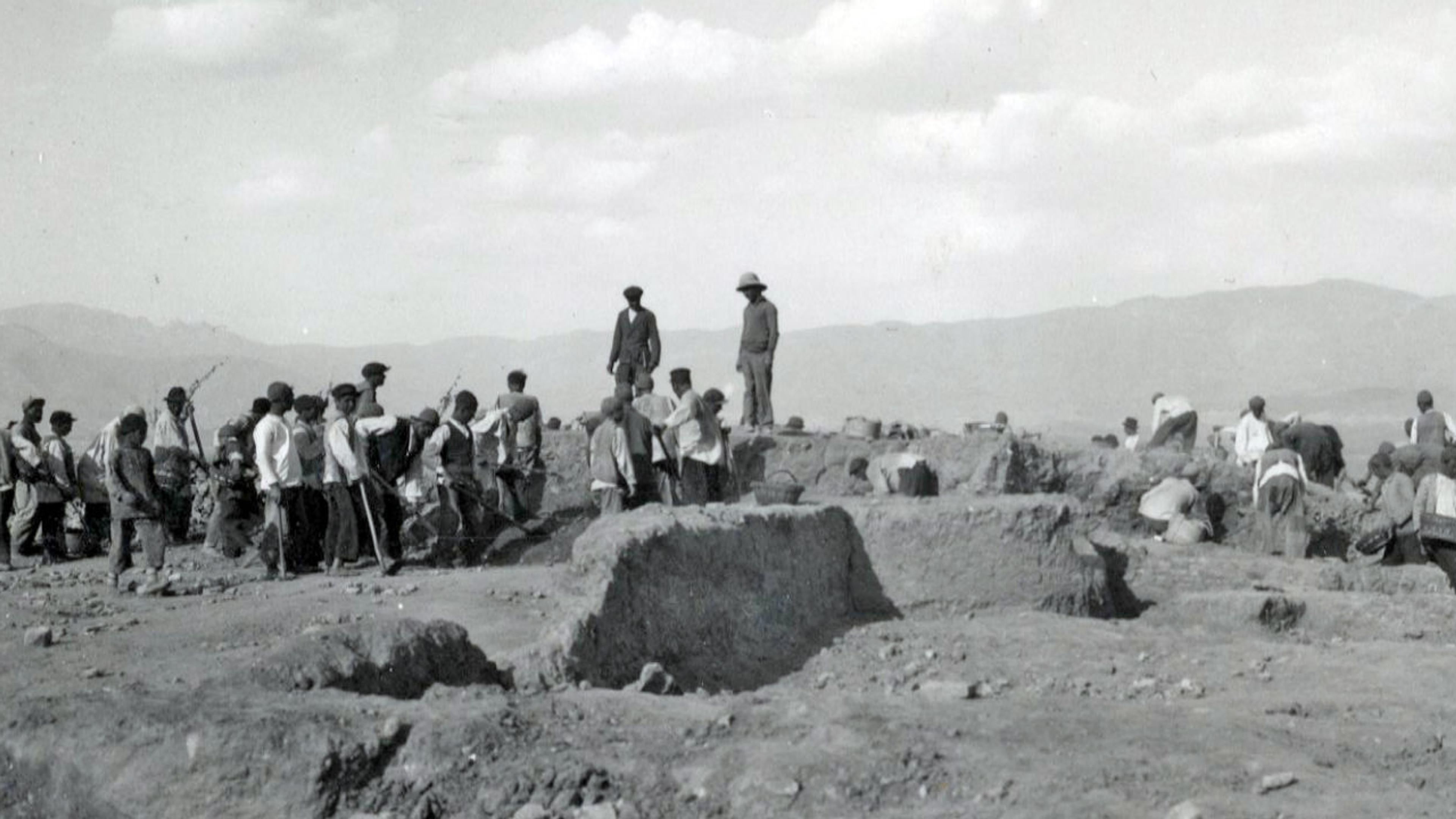 Archaeologists and laborers excavate an ancient site, surrounded by mountains under a cloudy sky, in a historical black-and-white photograph.