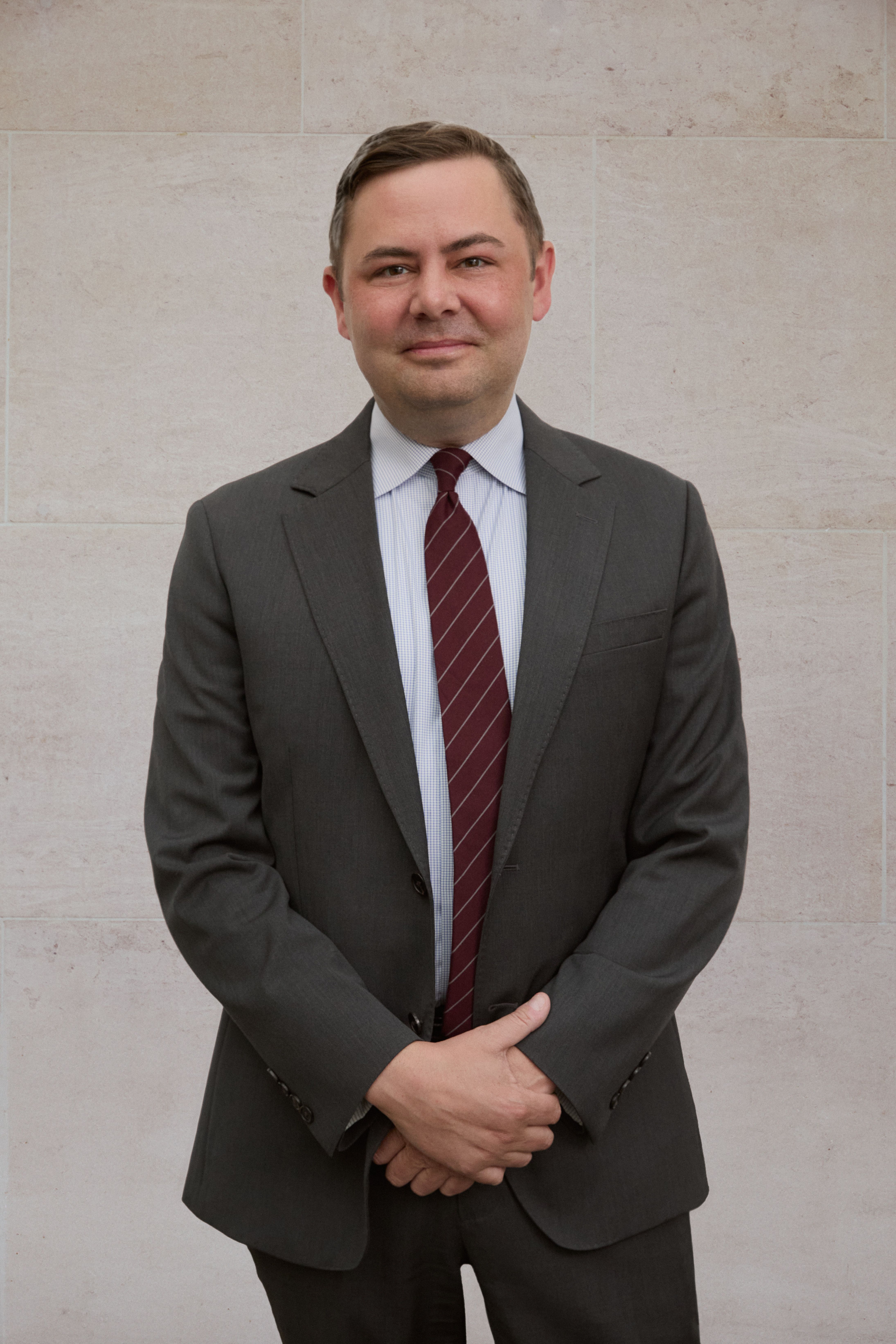 A color photograph of Paul Pineau standing in front of a stone wall in a dark gray suit with a red necktie.