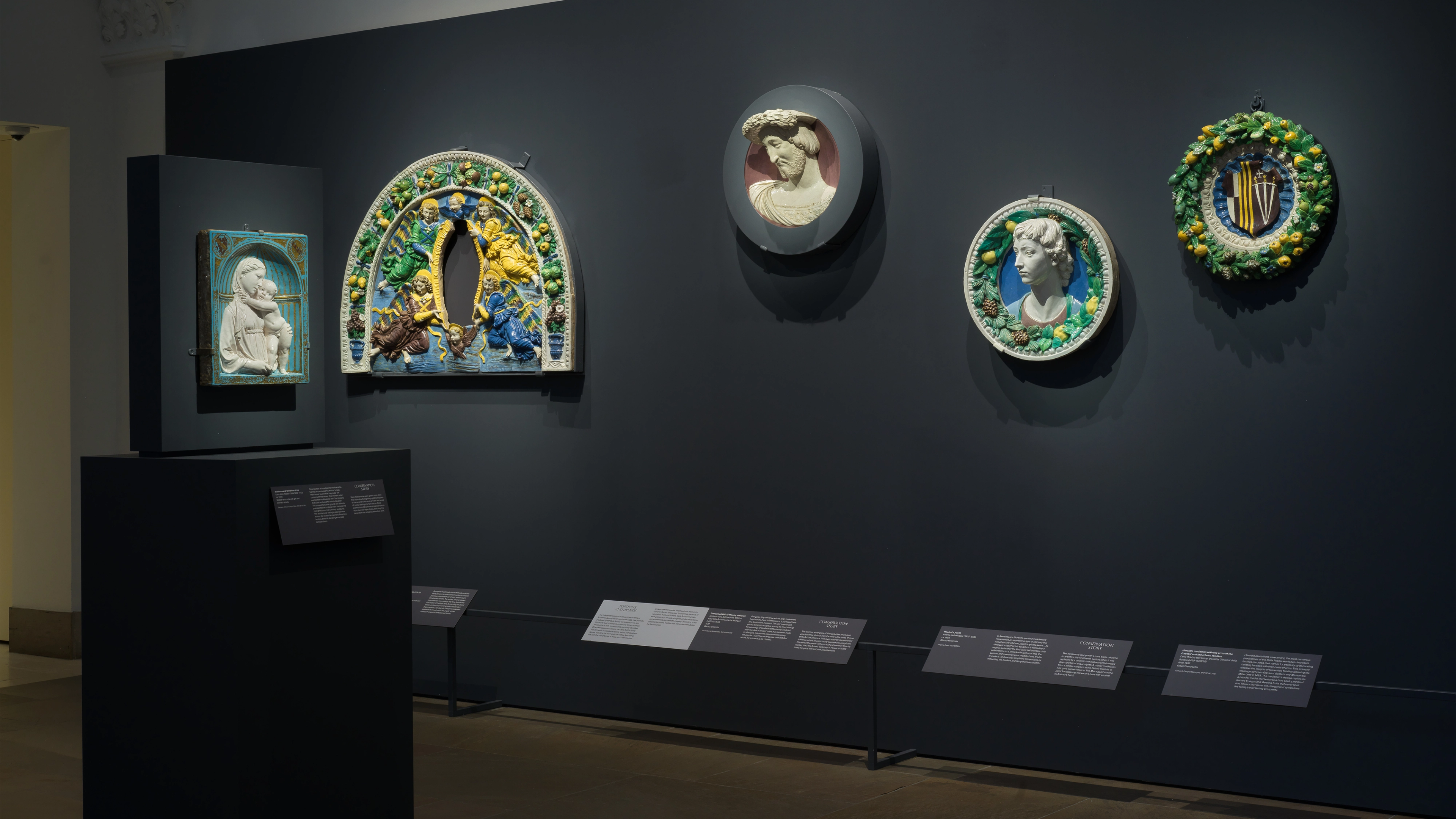A dim museum gallery displays five colorful framed ceramic reliefs on a charcoal wall, with small labels below