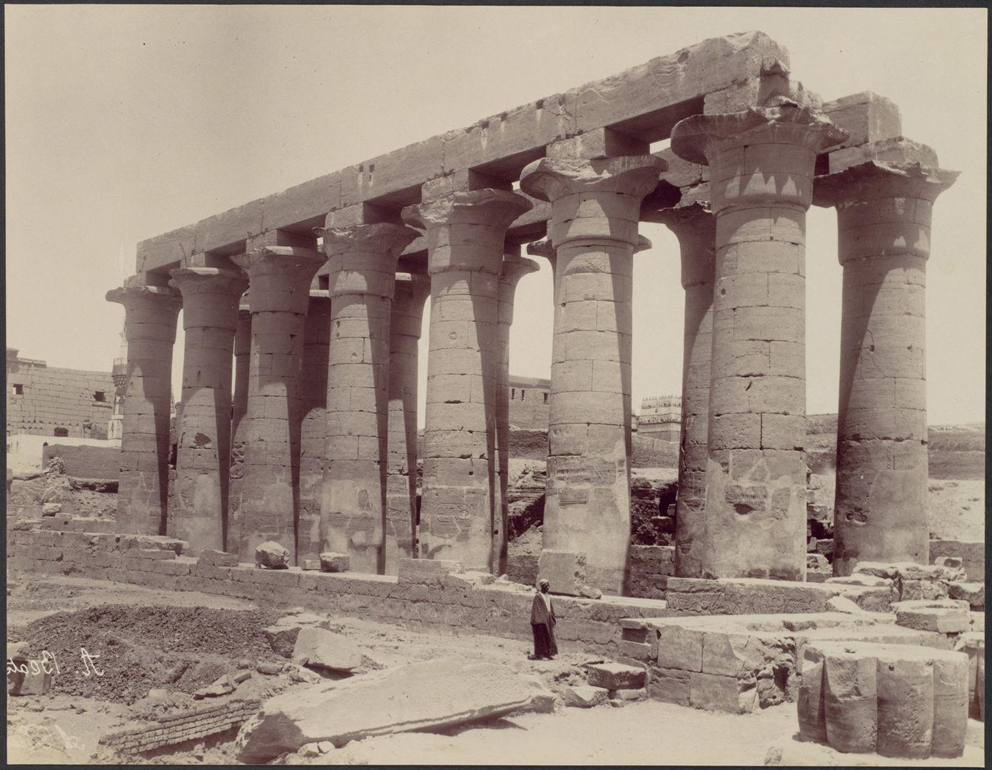 Black and white photo of a colonnade with two rows of massive columns topped by an architrave. The figure of a man in a long robe stands in the foreground.