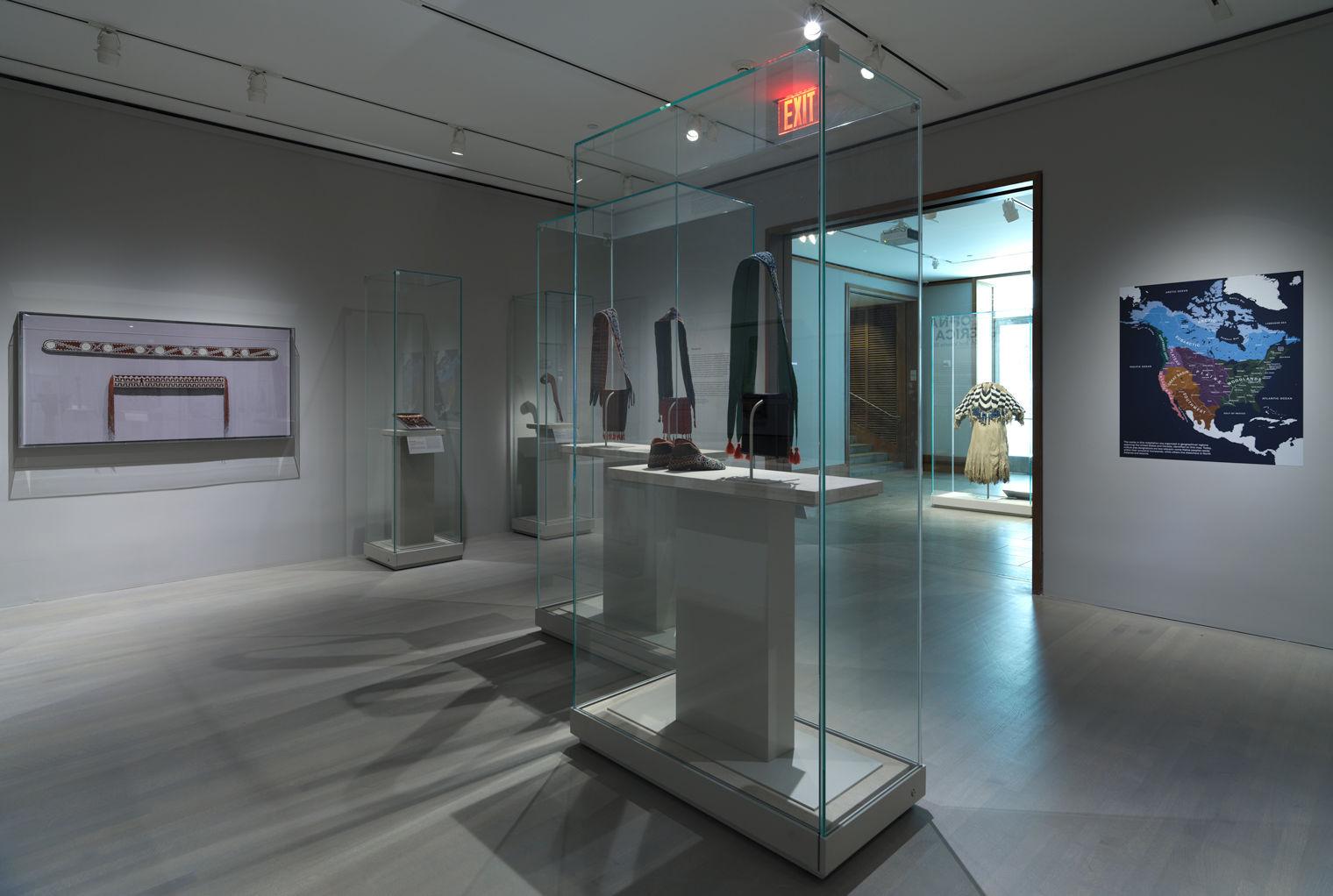 A wide view of the exhibition "Art of Native America" at The Met, featuring artworks from tribes in the Woodlands nation, including several shoulder bags in glass cases. On the right there is a map showing Native American territories in the United States.