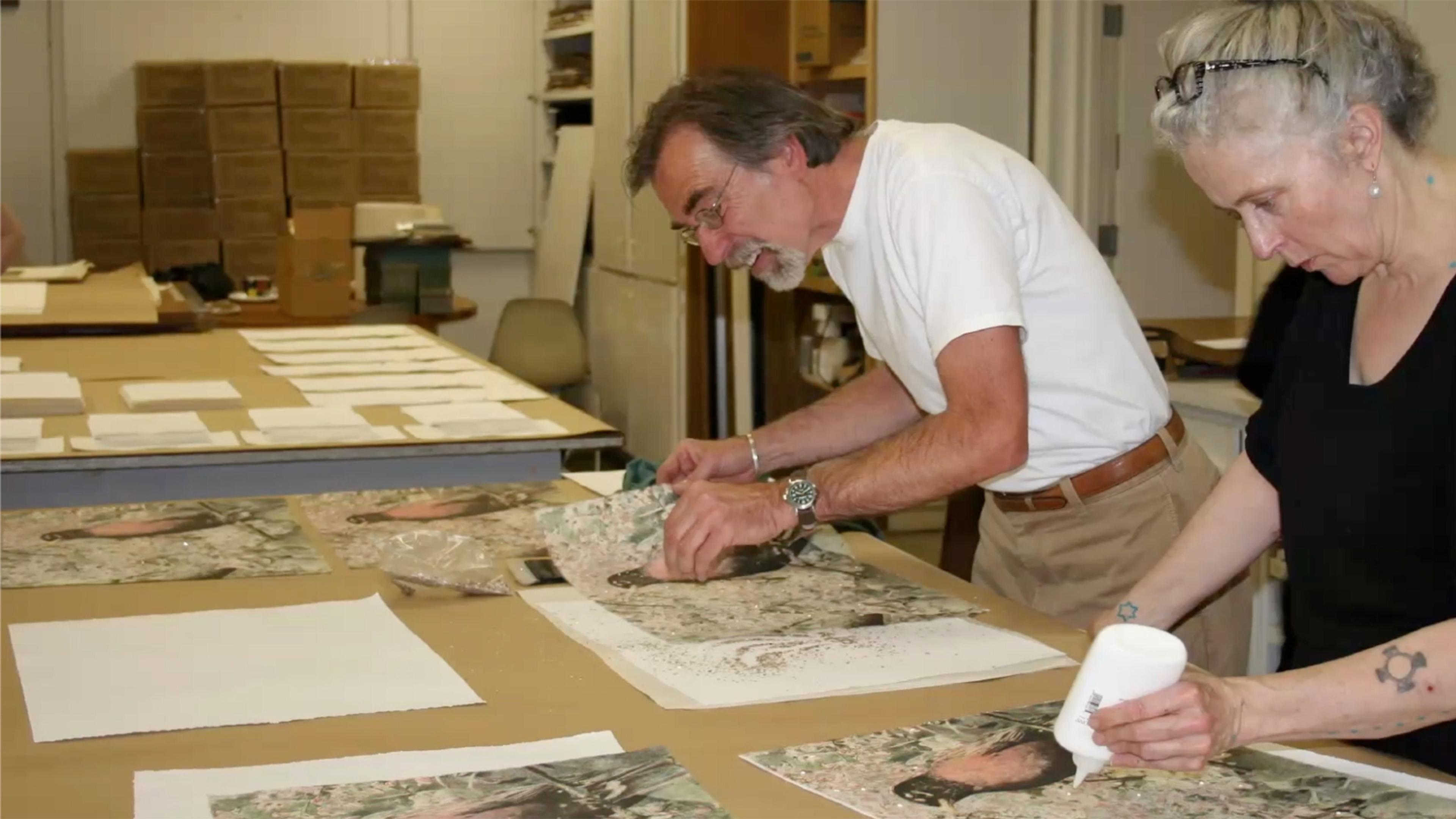 Two figures lean over a table and assemble paper-based artworks.