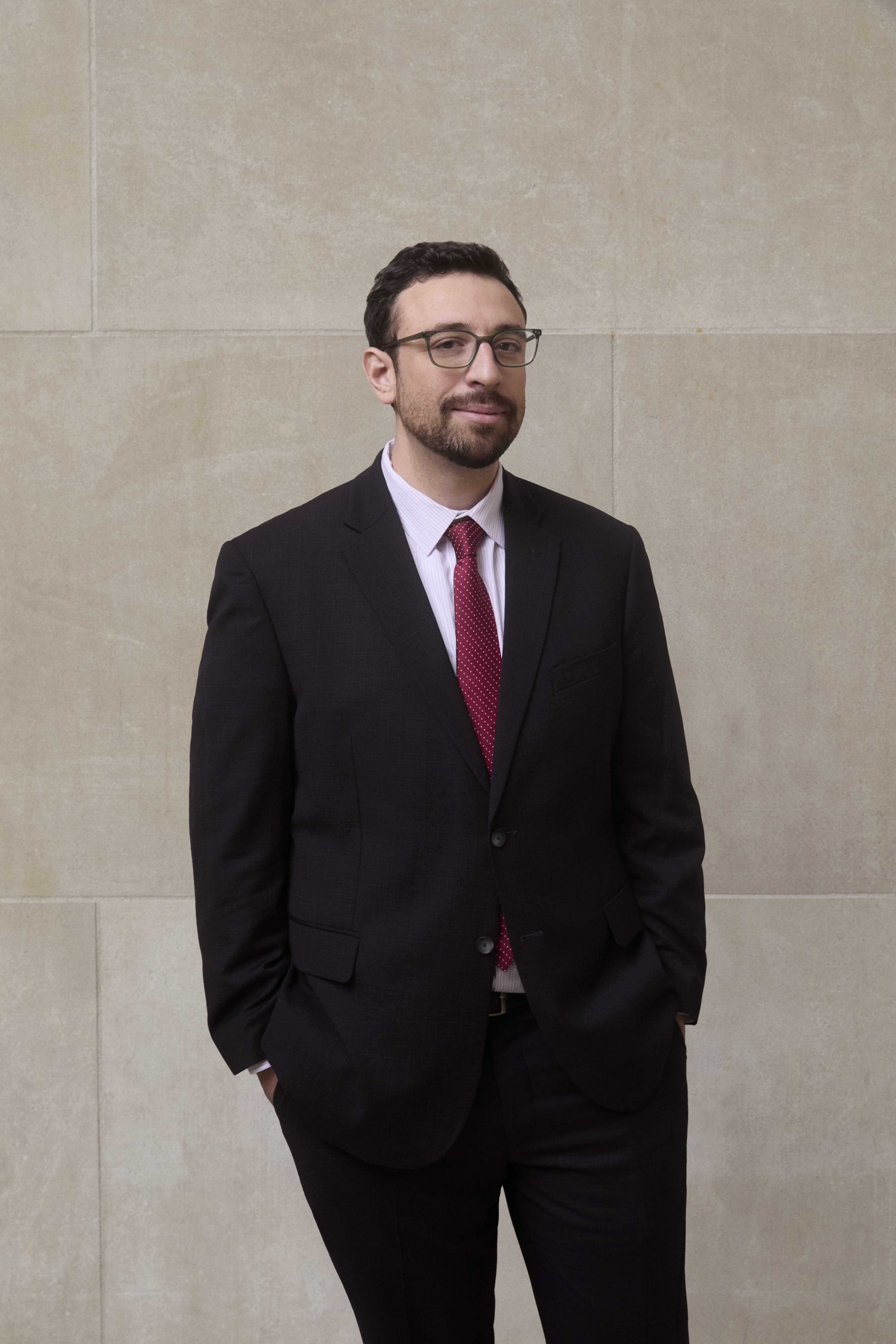 Color photograph of Dan Feeley standing in a dark suit with a white shirt and red tie. 