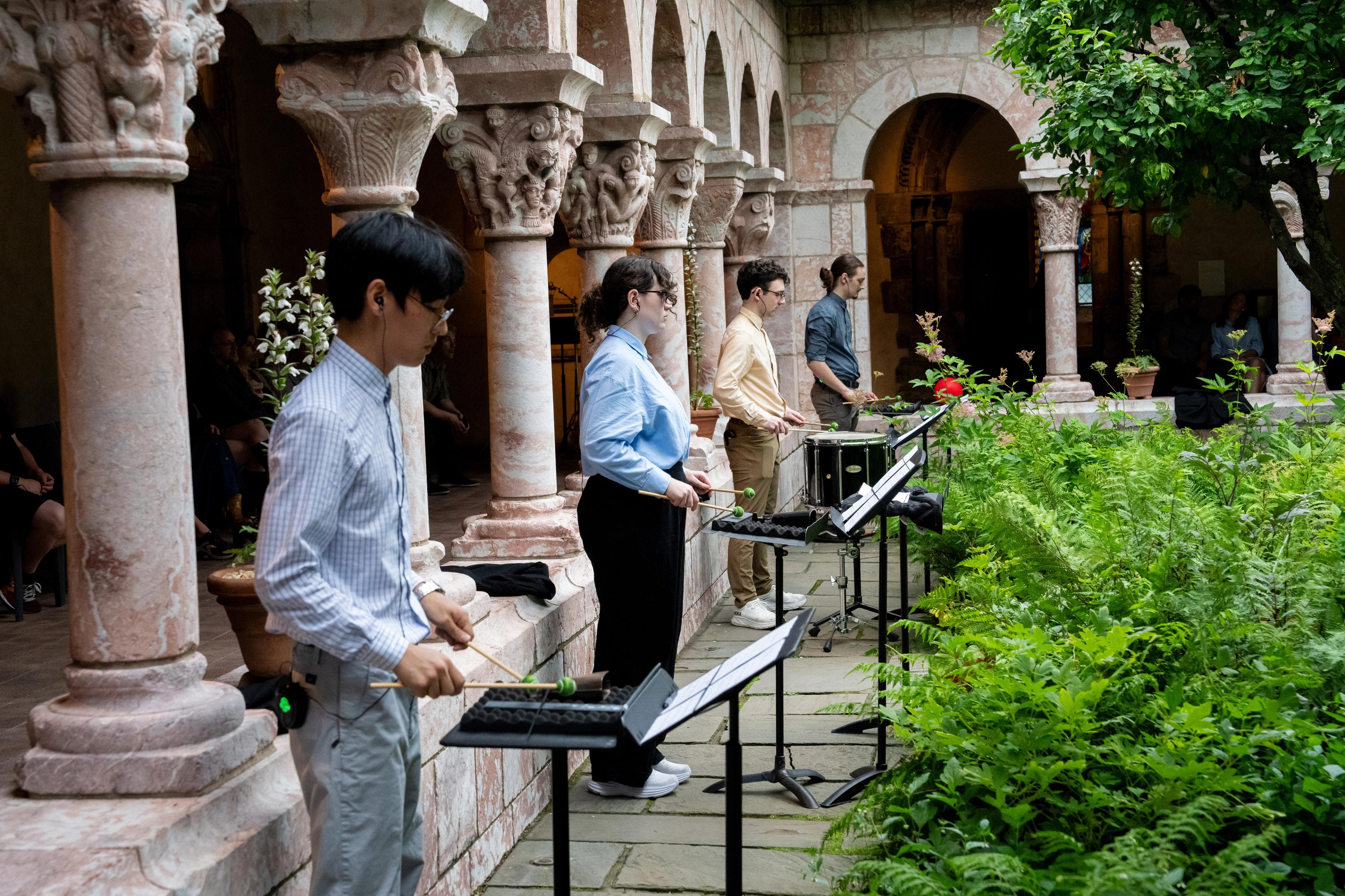 Four percussionists playing music between elegant stone pillars and verdant ferns.