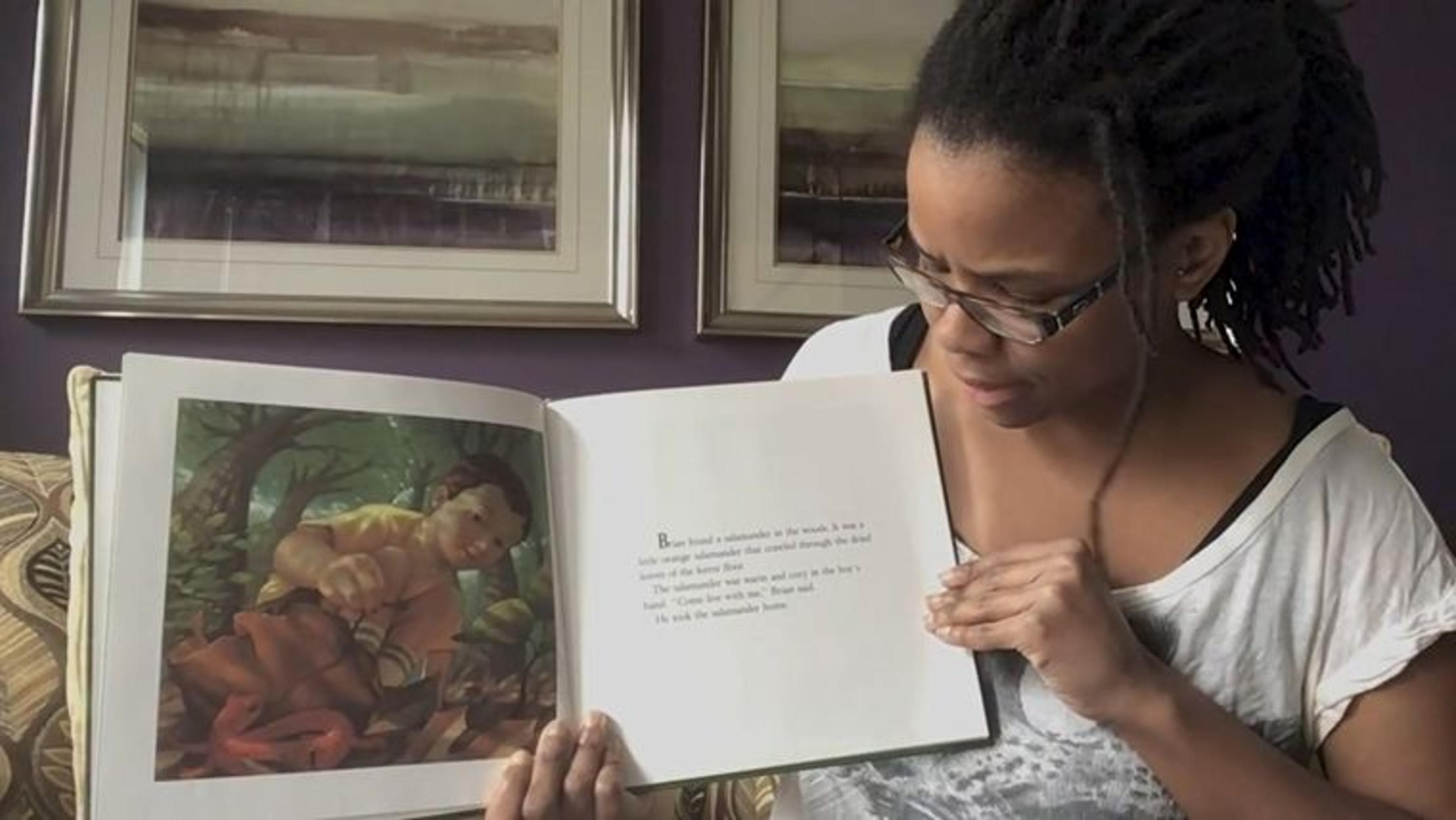 A woman holds a children's book towards a camera and looks at the page, reading.