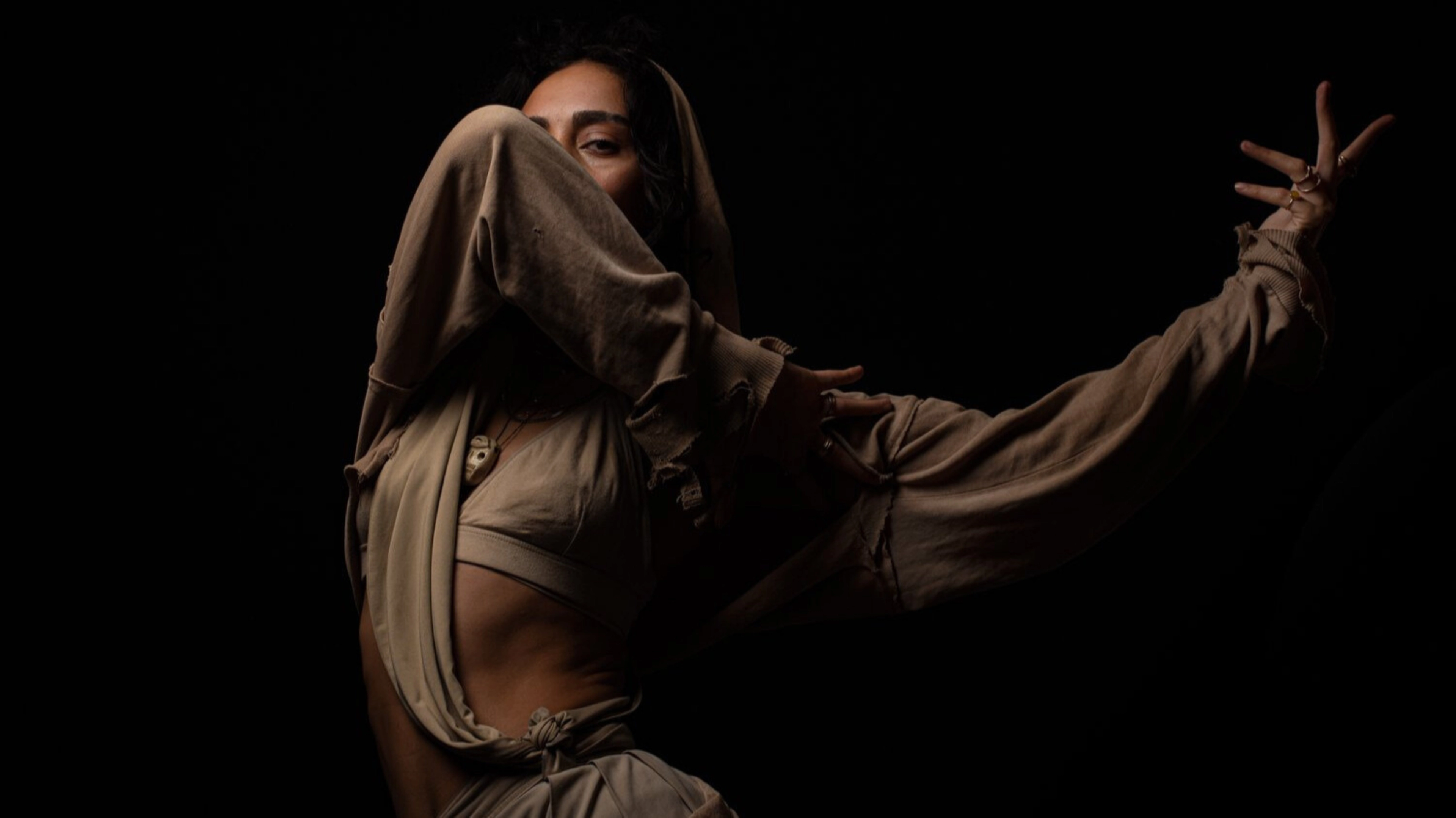 A dancer dressed in tan strikes a pose. Their elbow obscures their face and their left arm is outstretched to the side.