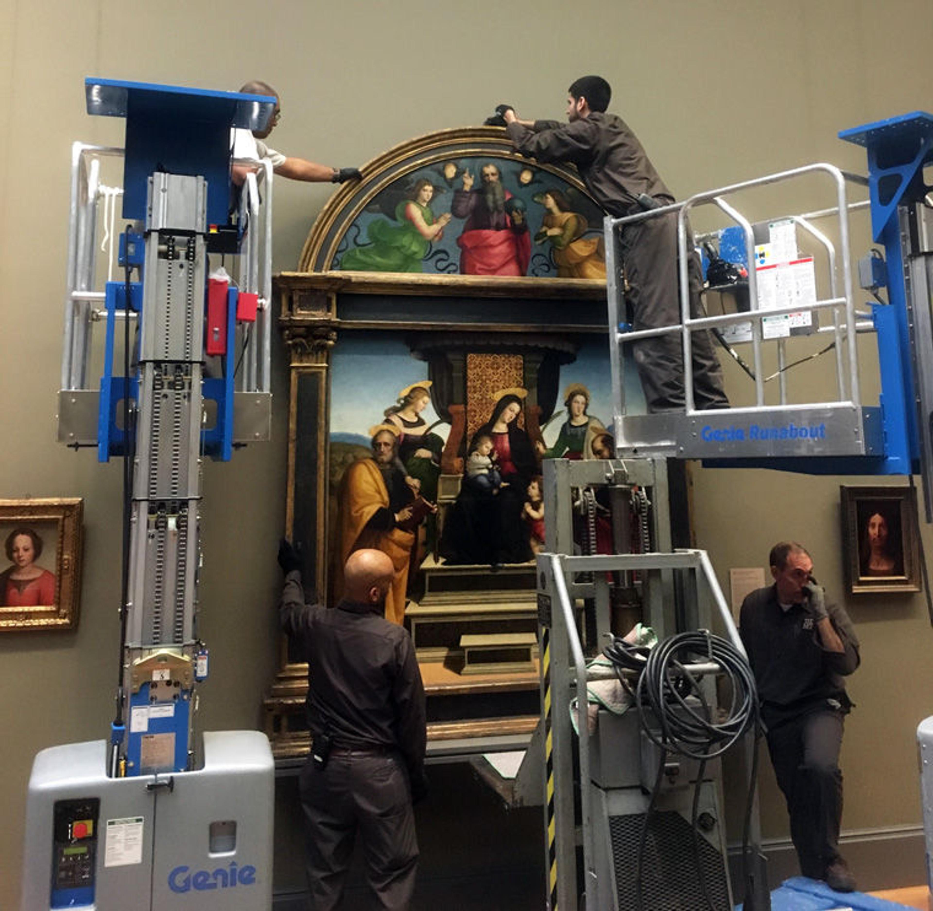 Workers using lifts to carefully install a large artwork on a museum wall, surrounded by smaller framed pieces.