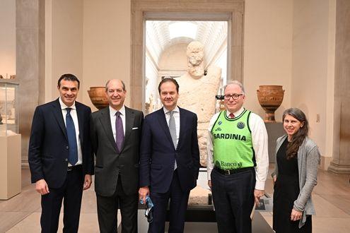A group photo with four men in suits and a woman in a long dress in front of a large stone sculpture of a man in a long light-filled gallery