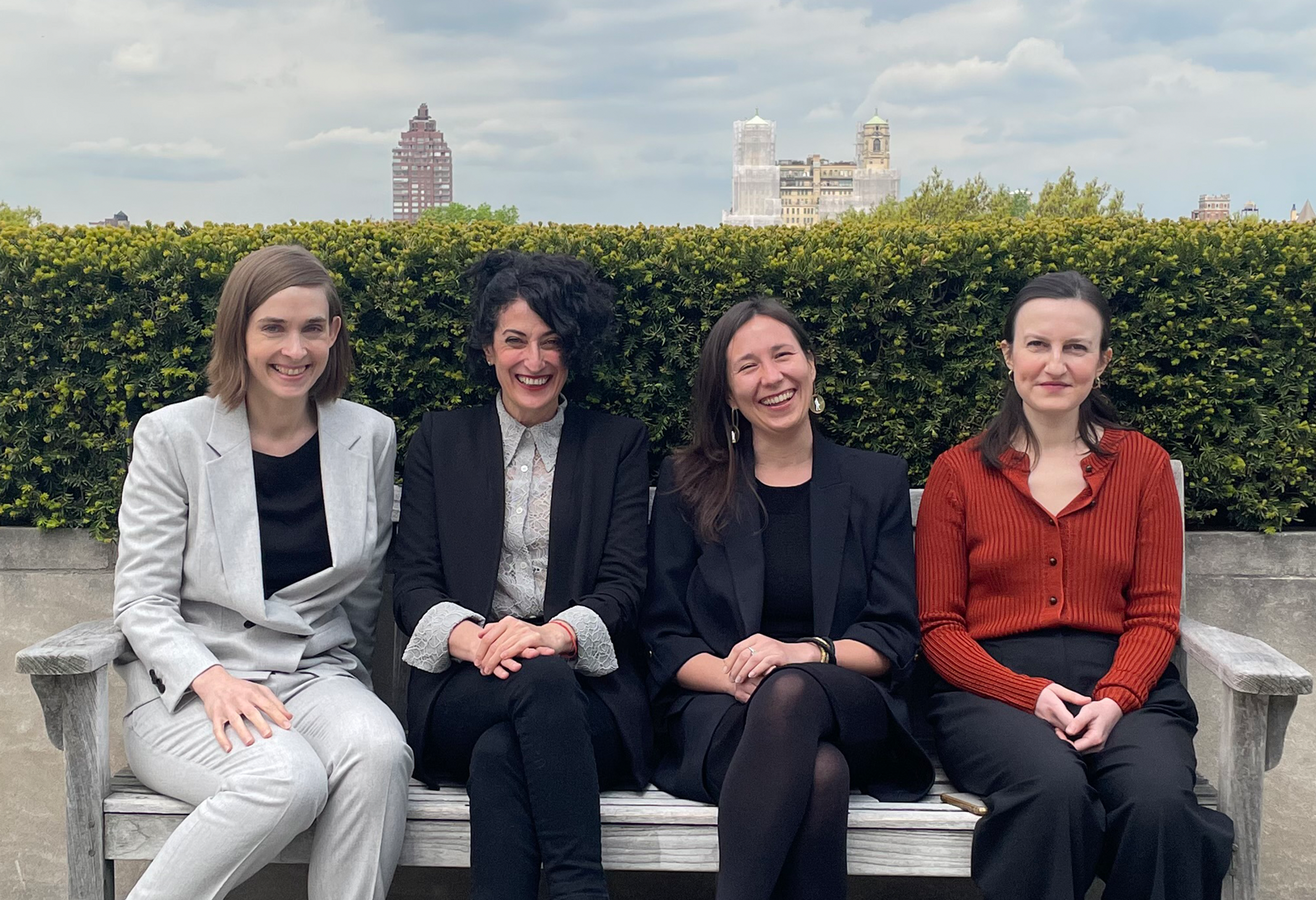 Four current fellows smiling and sitting on the roof of the Met with shrubbery and part of the city skyline in the background