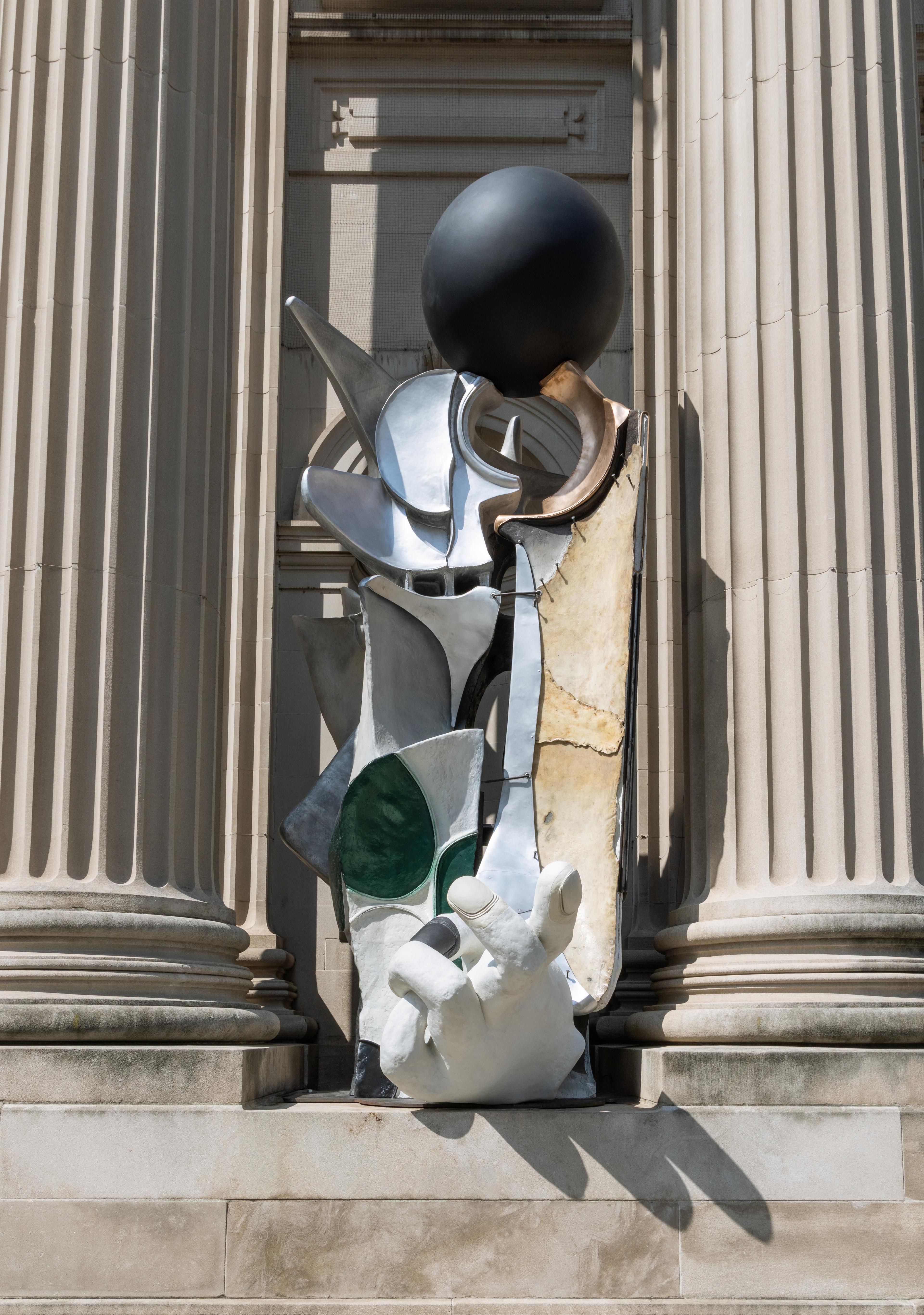 An abstract, totem-like sculpture standing in a niche between two fluted Neoclassical columns on a museum facade