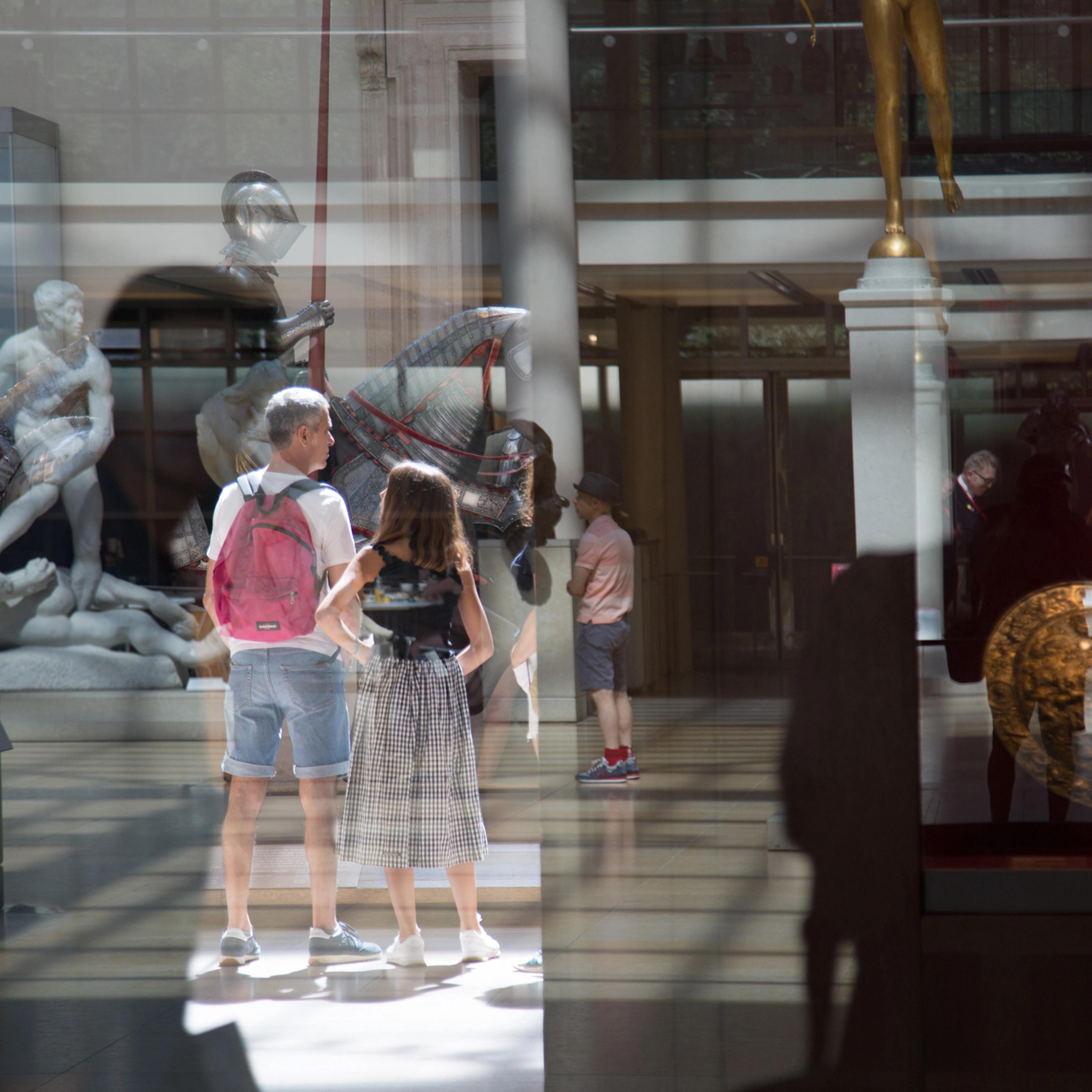 Photograph of reflecting stone sculptures, medieval armor, a round shield, and Museum patrons  in the sunlit Engelhard Court.