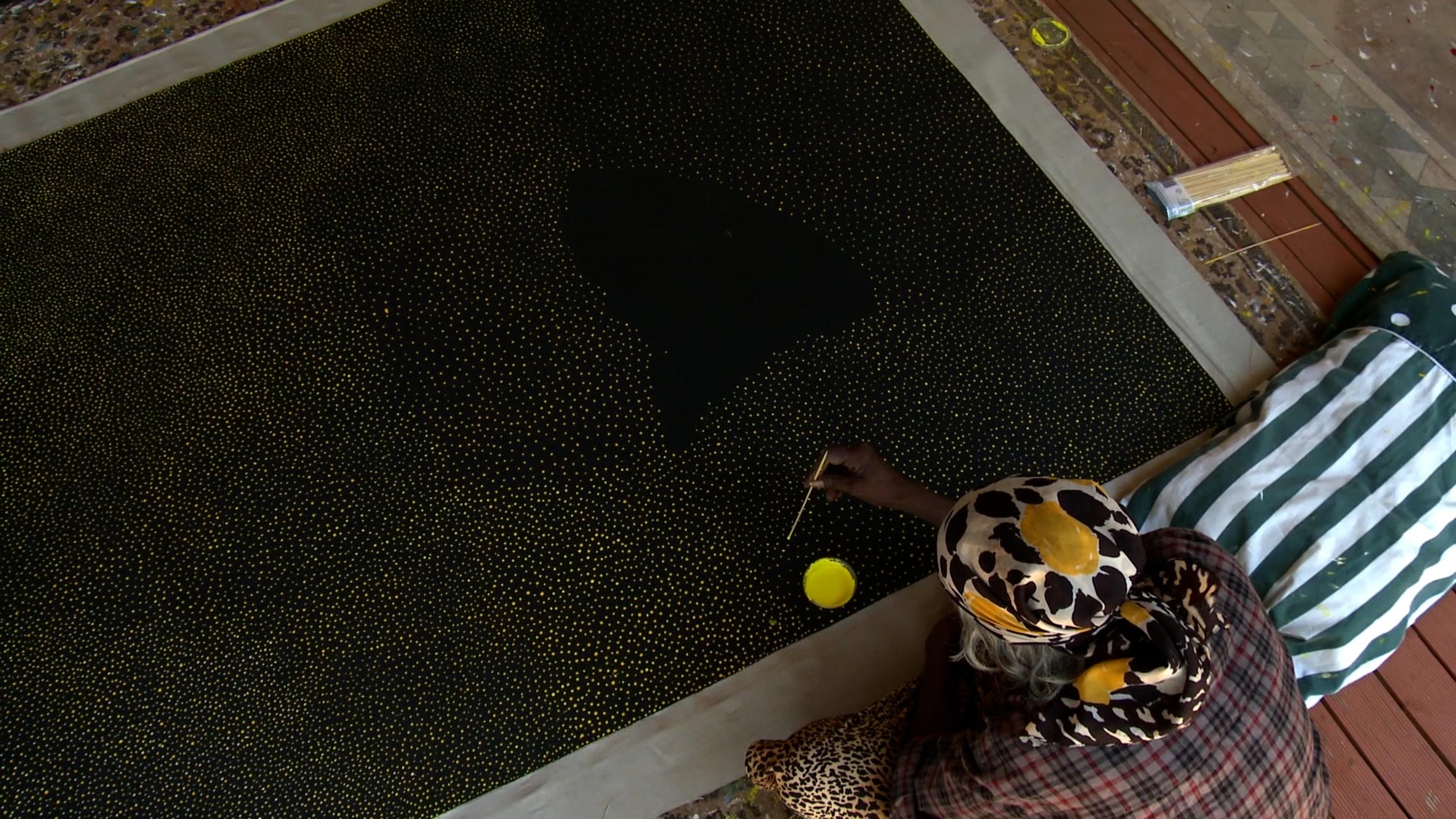 Woman painting yellow dots across a black canvas laid on the ground