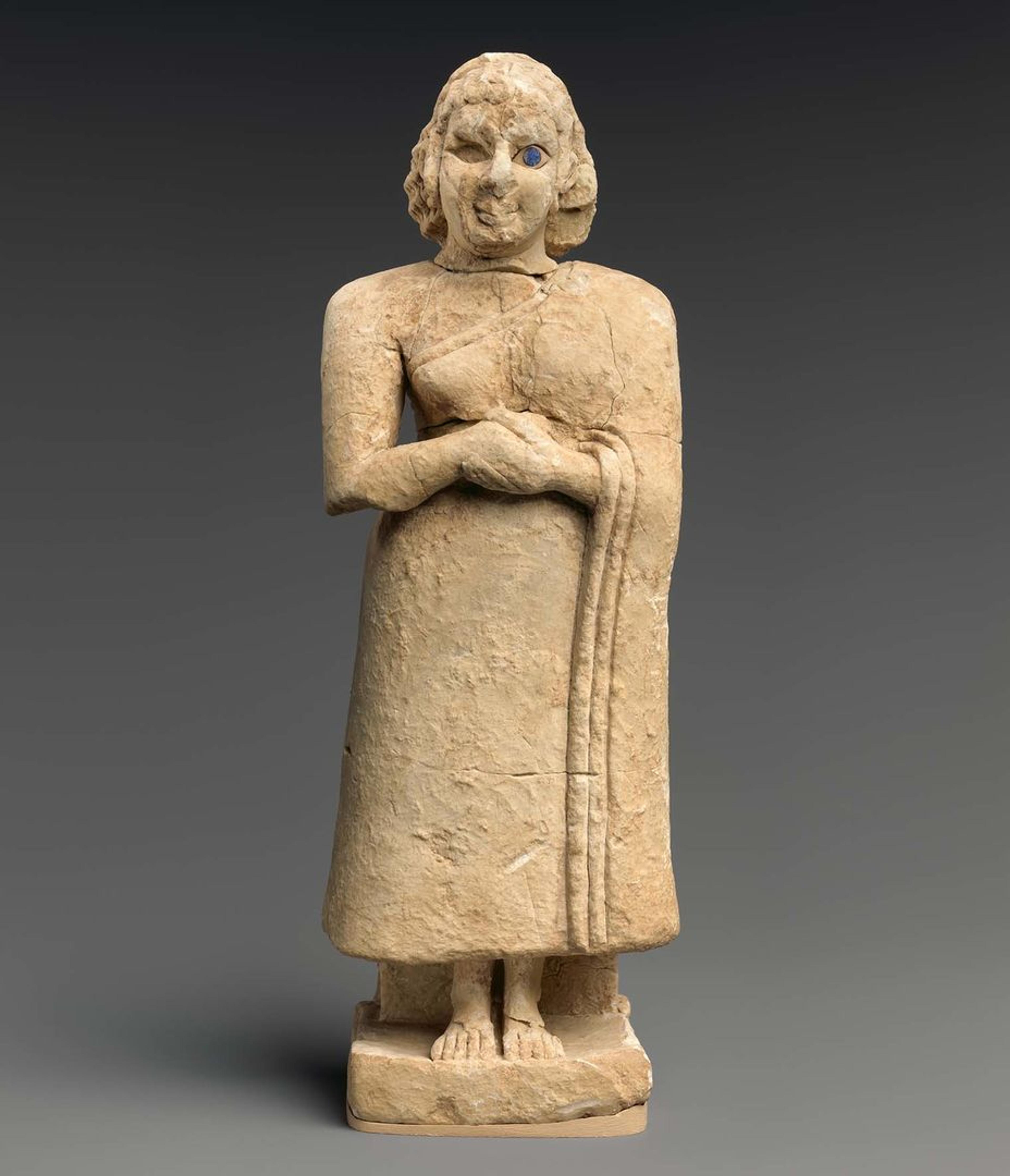 A limestone sculpture of a woman standing with hands clasped in prayer. Inlaid with shell and lapis lazuli for details.