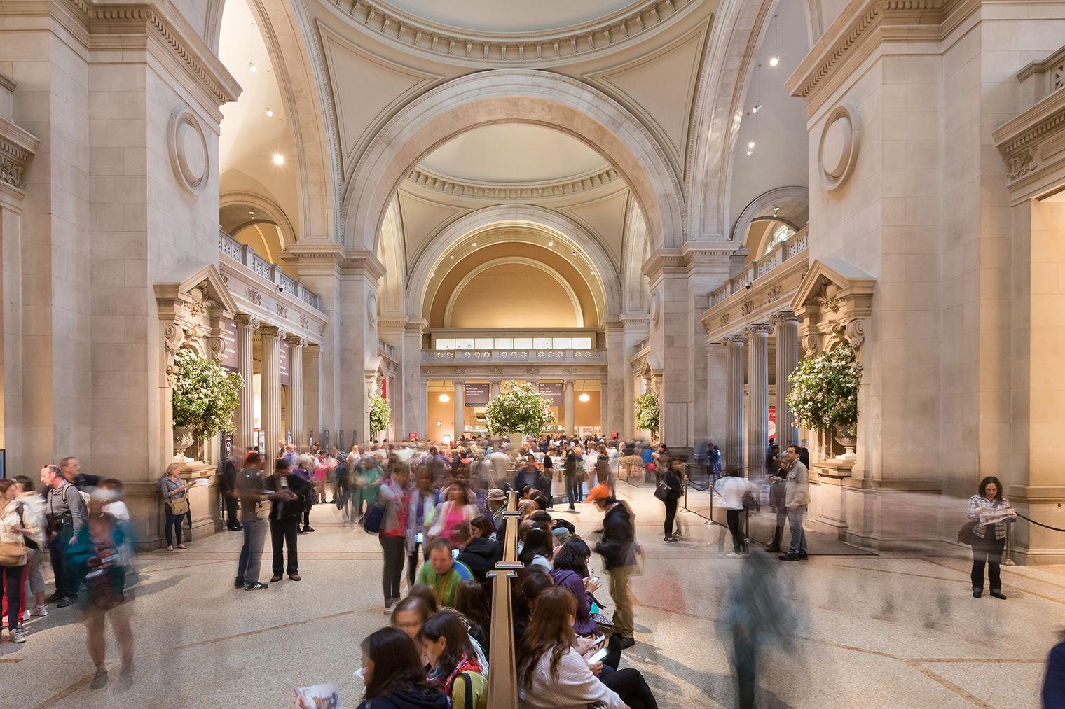 A bustling photo: people are gathered at the information desk, looking at maps, and resting in seats, against the majestic columns and domes of The Met's Great Hall.