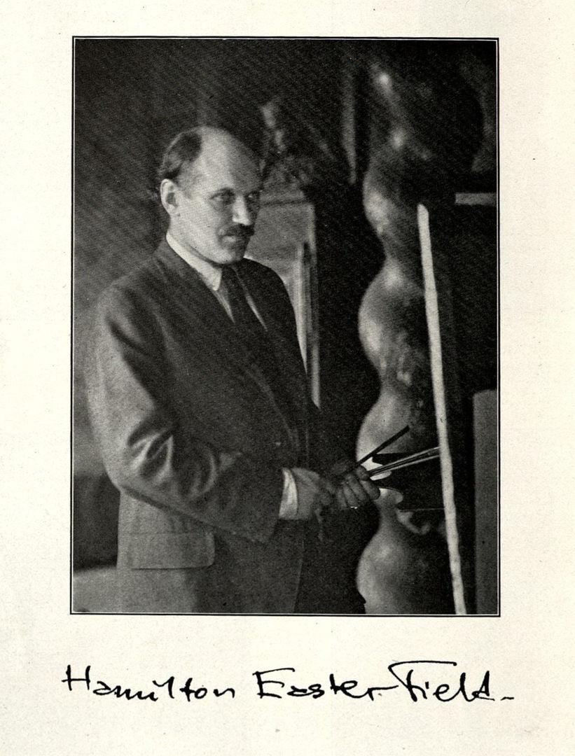 Black and white photograph of a white man in a suit with dark hair and a mustache. He his holding a bunch of paint brushes in front of a canvas. Below, there is a signature that says Hamilton Easter Field.