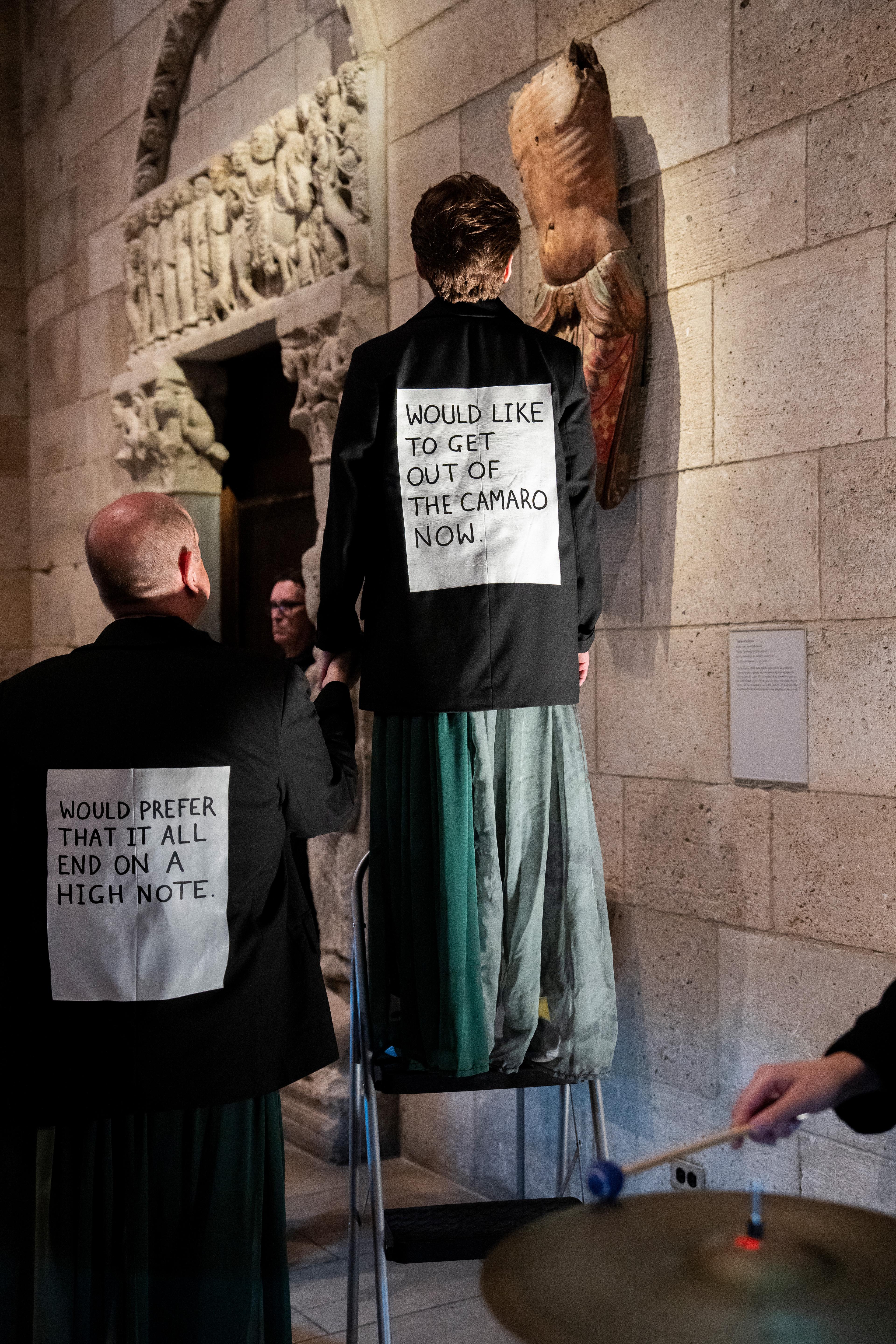 Two performers stand with their backs to the viewer. The backs of their jackets have white cards with whimsical phrases.