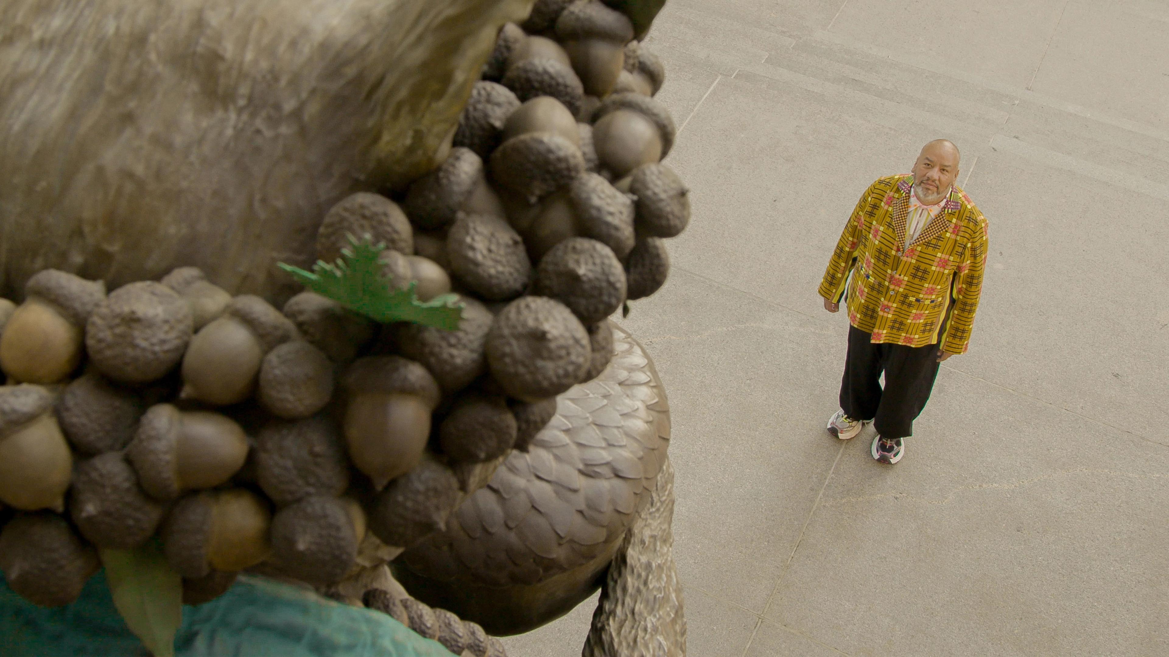 A bald man in a colorful plaid jacket looks up at a large sculpture adorned with acorns. The setting is an urban plaza, conveying curiosity and contemplation.