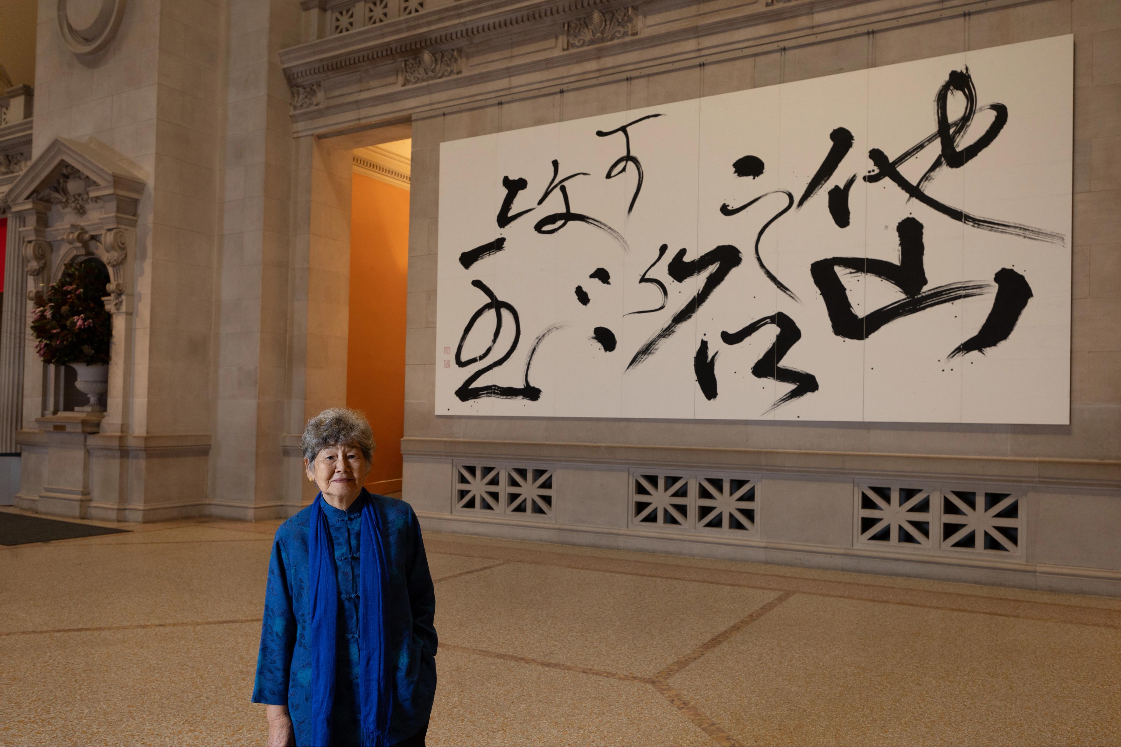Woman wearing all blue standing tall, and smiling with a piece of art calligraphy in the back.