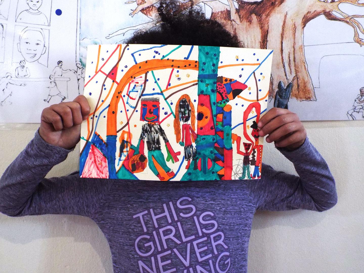 Awra holds her finished art up high in front of her face. In it are abstract figurines in bright pink, orange, and turquoise in a geometric setting.