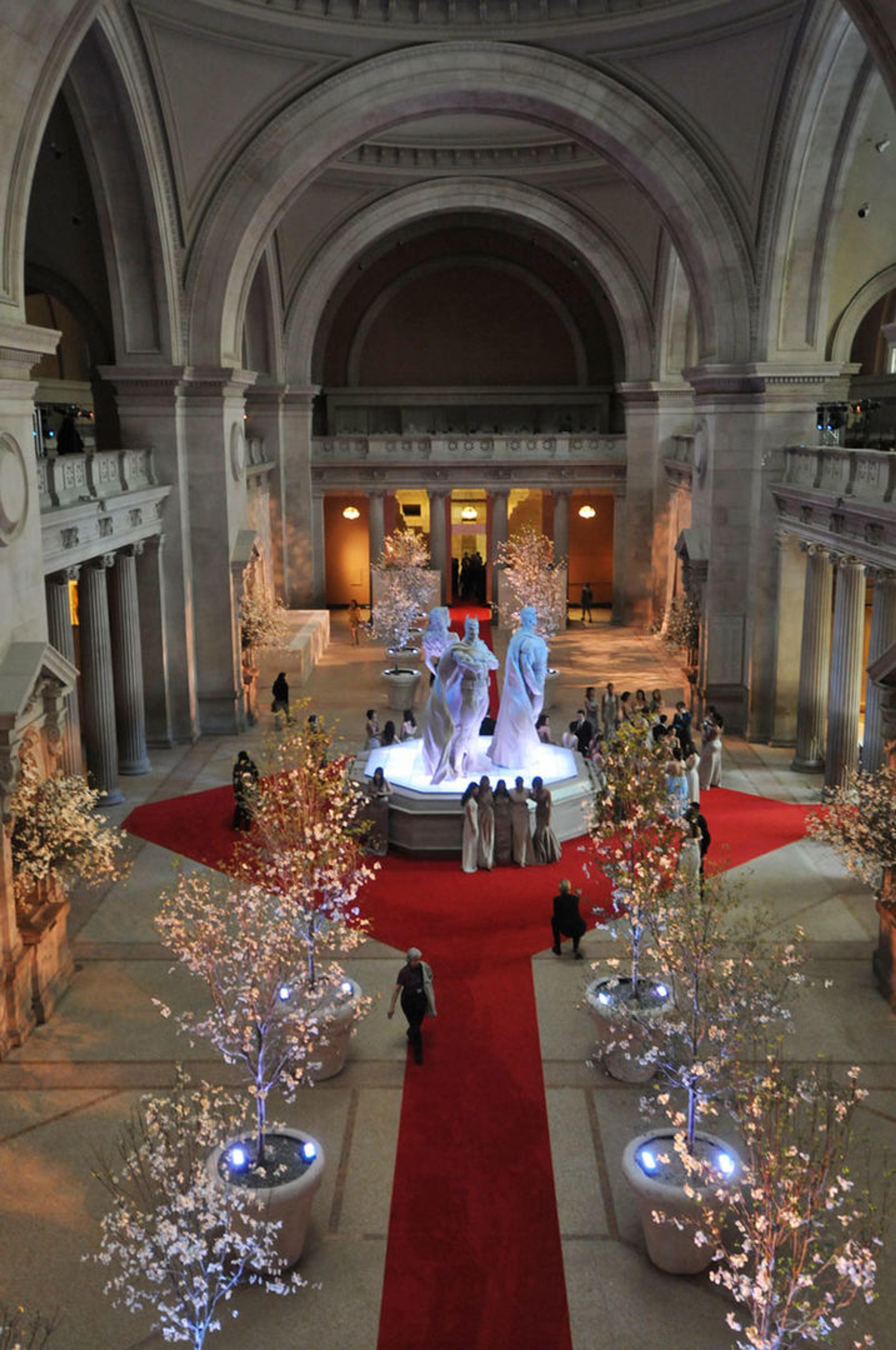 A view of The Met's Great Hall during the 2008 Met Gala, featuring large superhero sculptures atop an information desk and many elegantly lit trees
