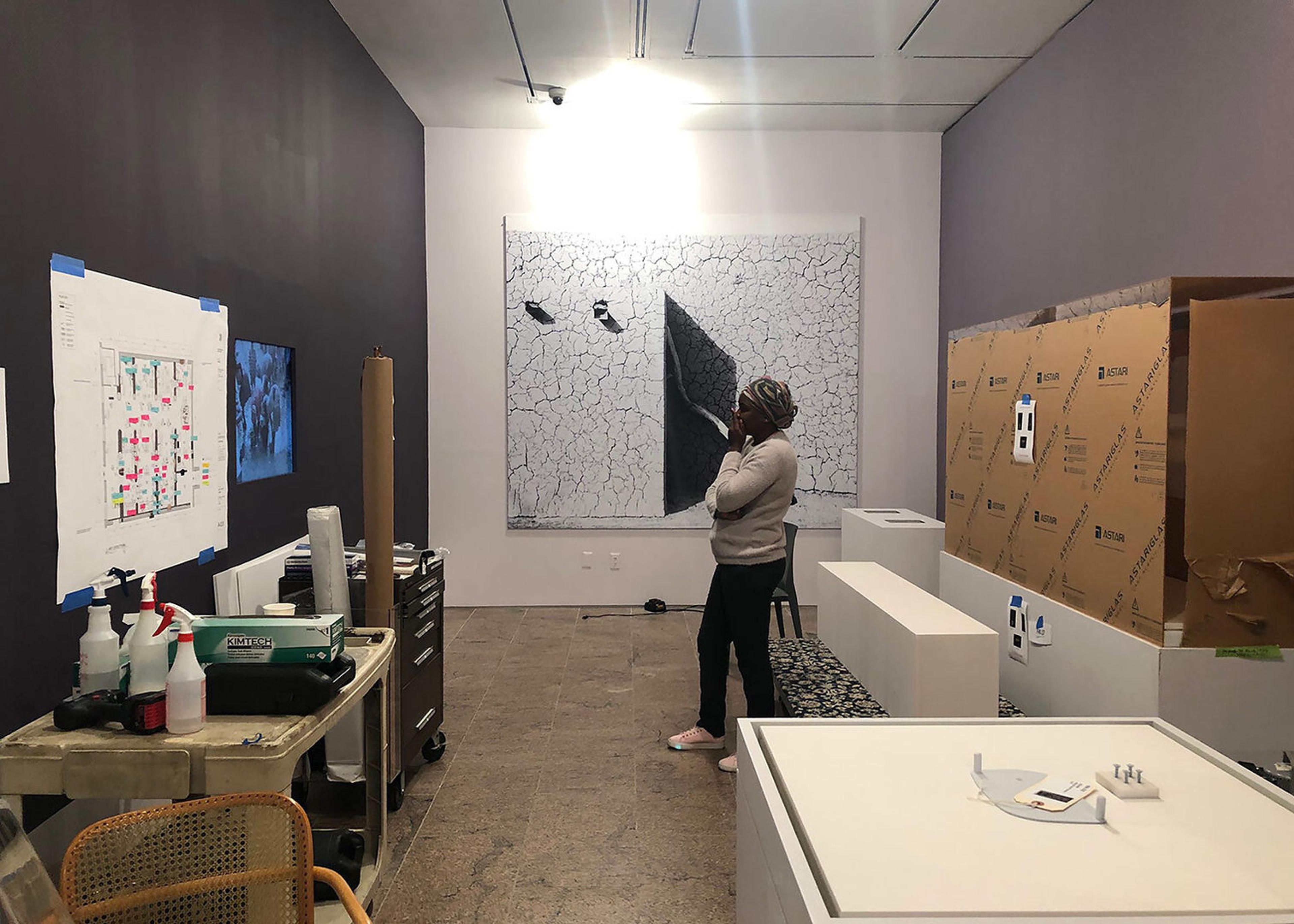 A woman stands in a brightly lit room with a large cardboard box, various white containers, and a large white floor plan hanging on the wall