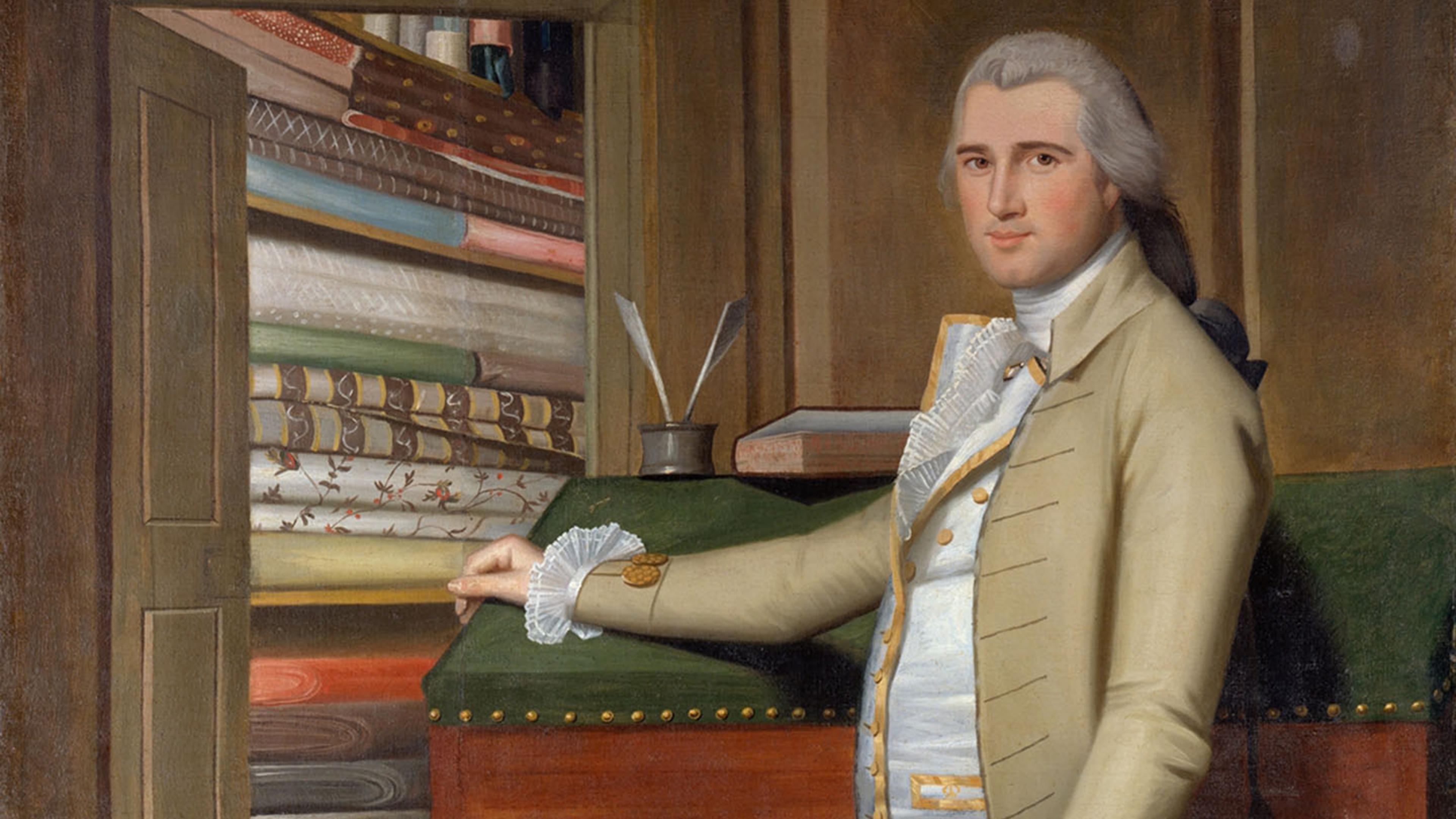 A man dressed in colonial attire rests his hand on a writing desk adjacent to an open door that looks into a room of shelves storing an abundance of folded textiles