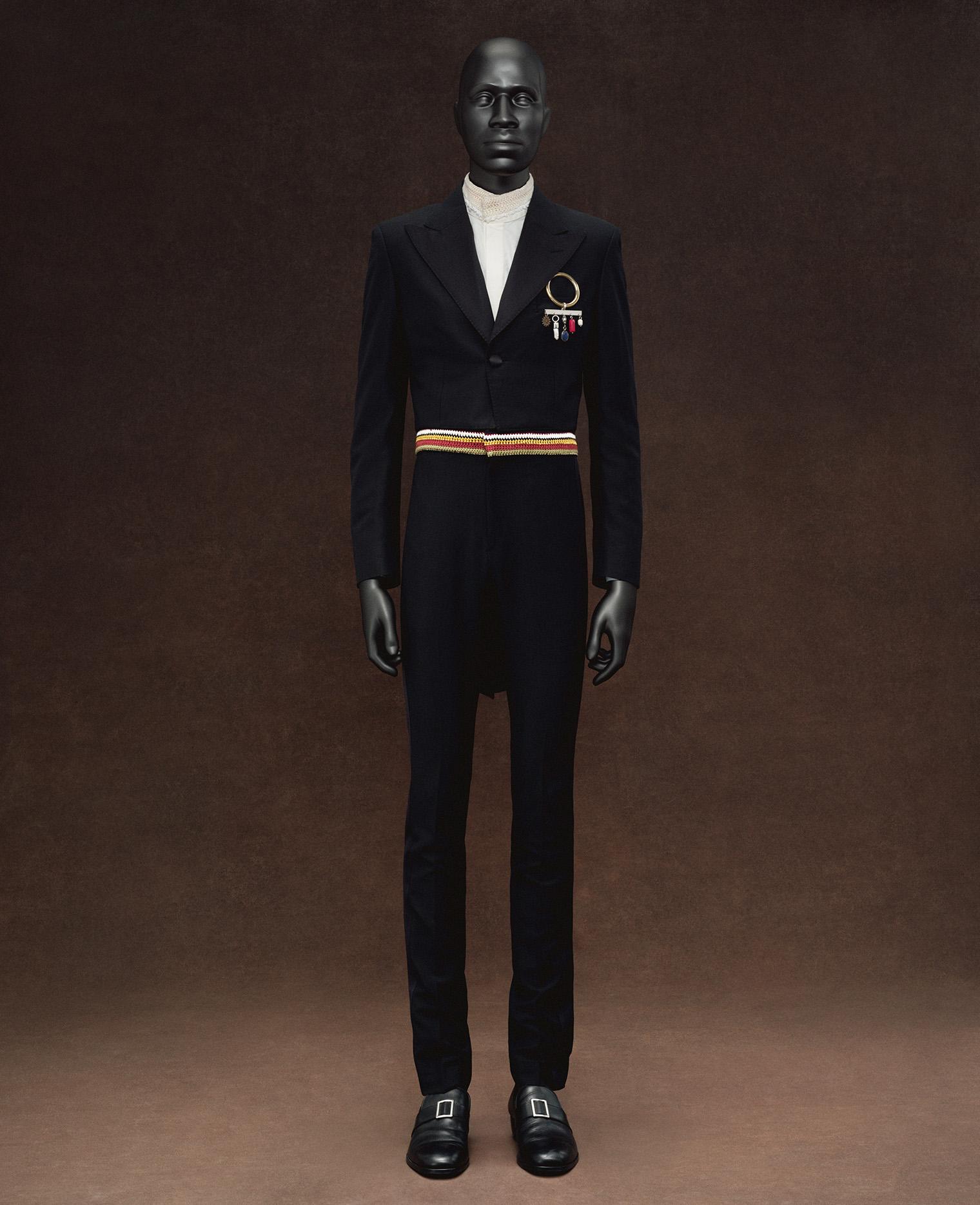 A mannequin is dressed in an elegant black suit with a high-neck white shirt, adorned with colorful military-style medals and a red-striped belt.