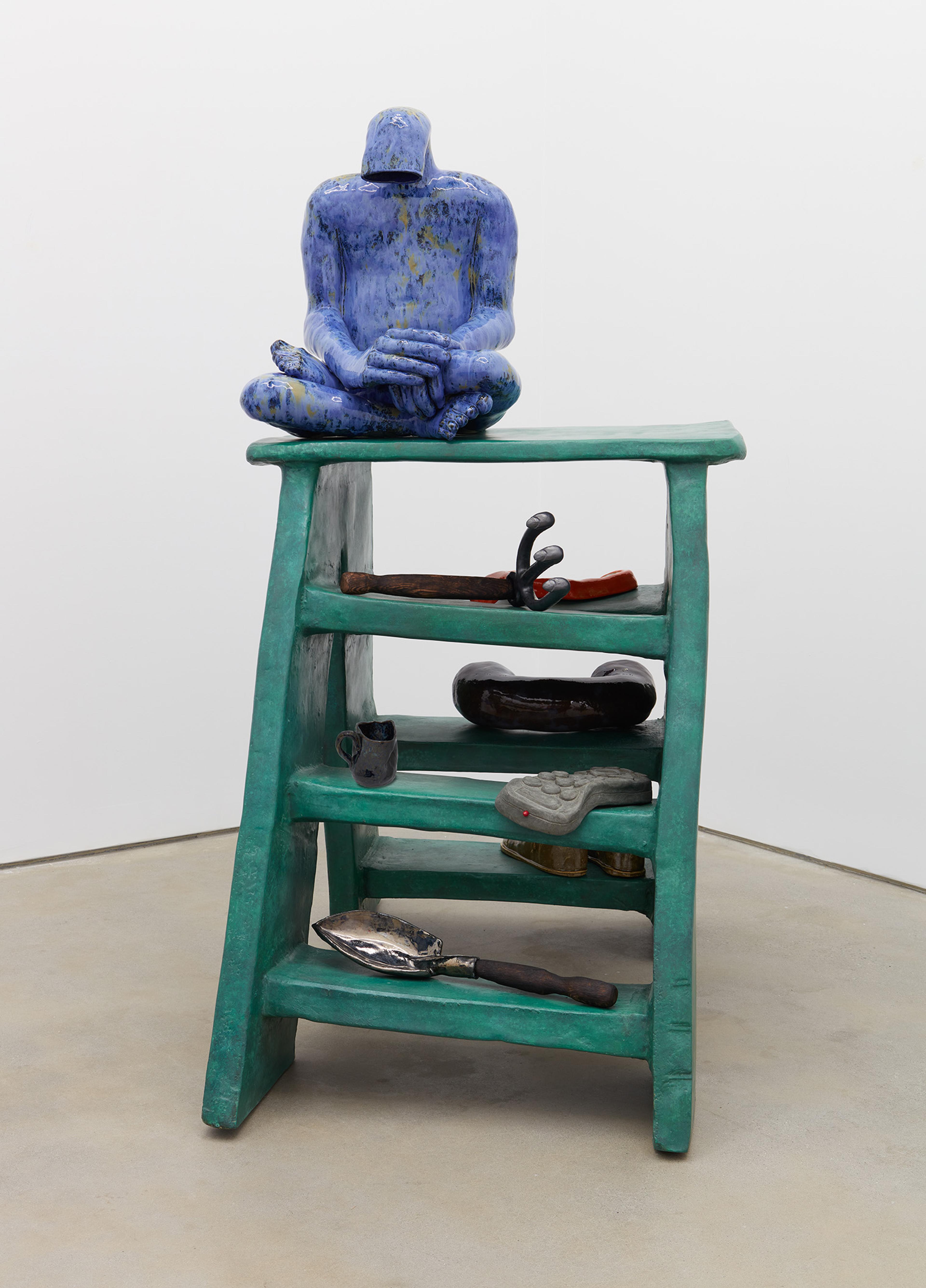 A sculpted blue figure sits cross-legged atop a green, wavy wooden shelf. Below, various abstract objects rest on the shelves
