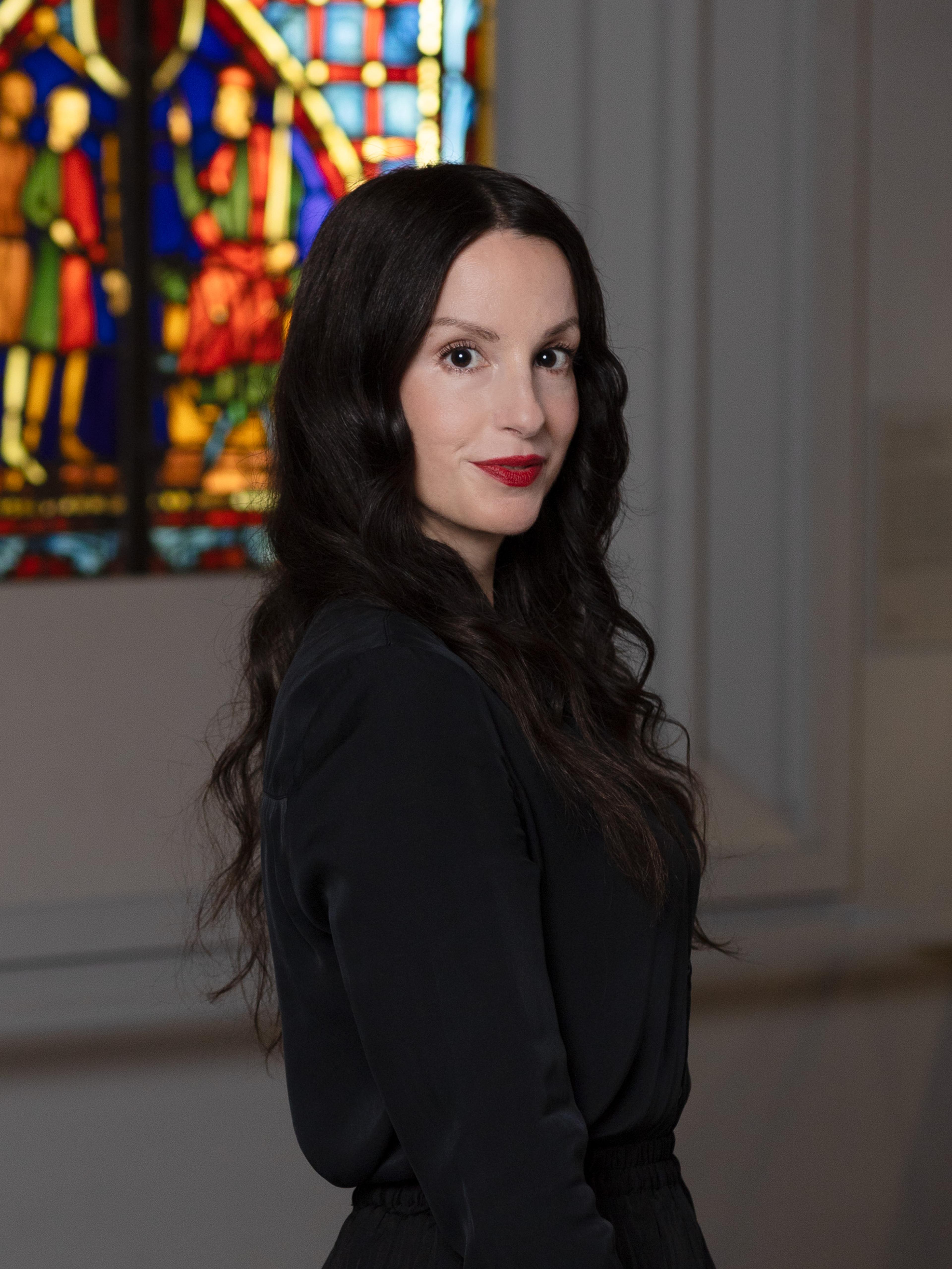 A woman with long brown hair smiles in front of a stained glass window