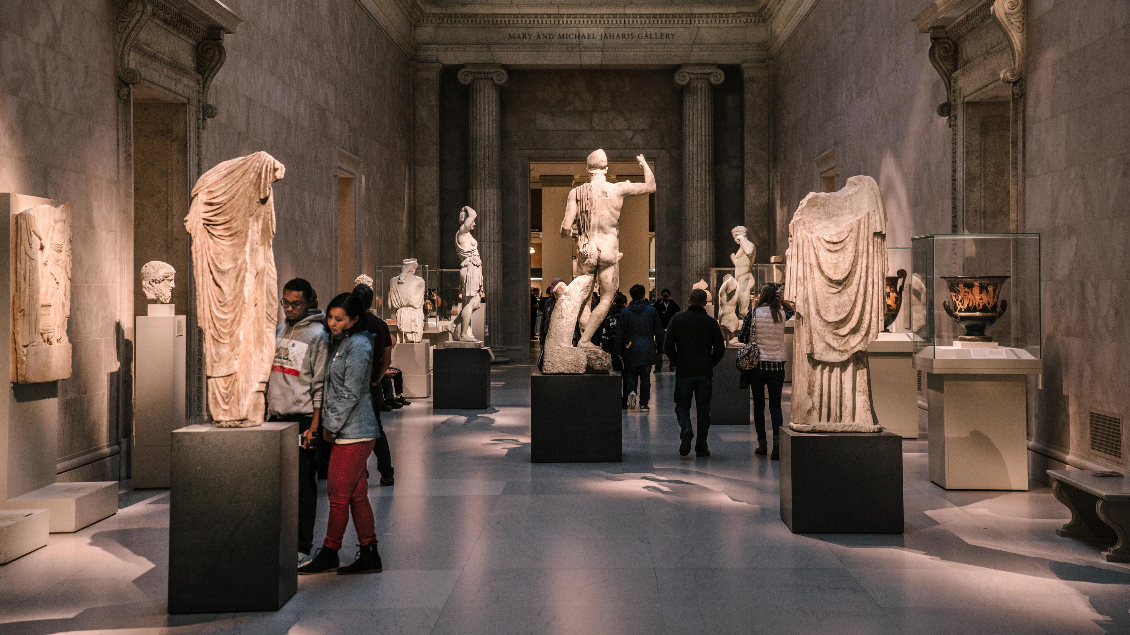 A gallery of white marble statues of human figures missing limbs and heads in art gallery with dramatic lighting. 