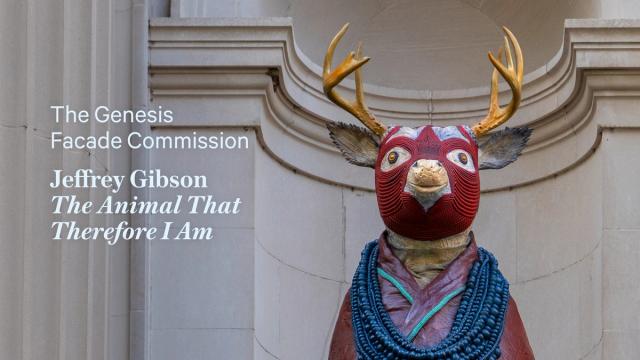 A statue of a deer with a red tribal mask and antlers stands in front of an ornate stone facade. Text reads “The Genesis Facade Commission, Jeffrey Gibson, The Animal That Therefore I Am.”
