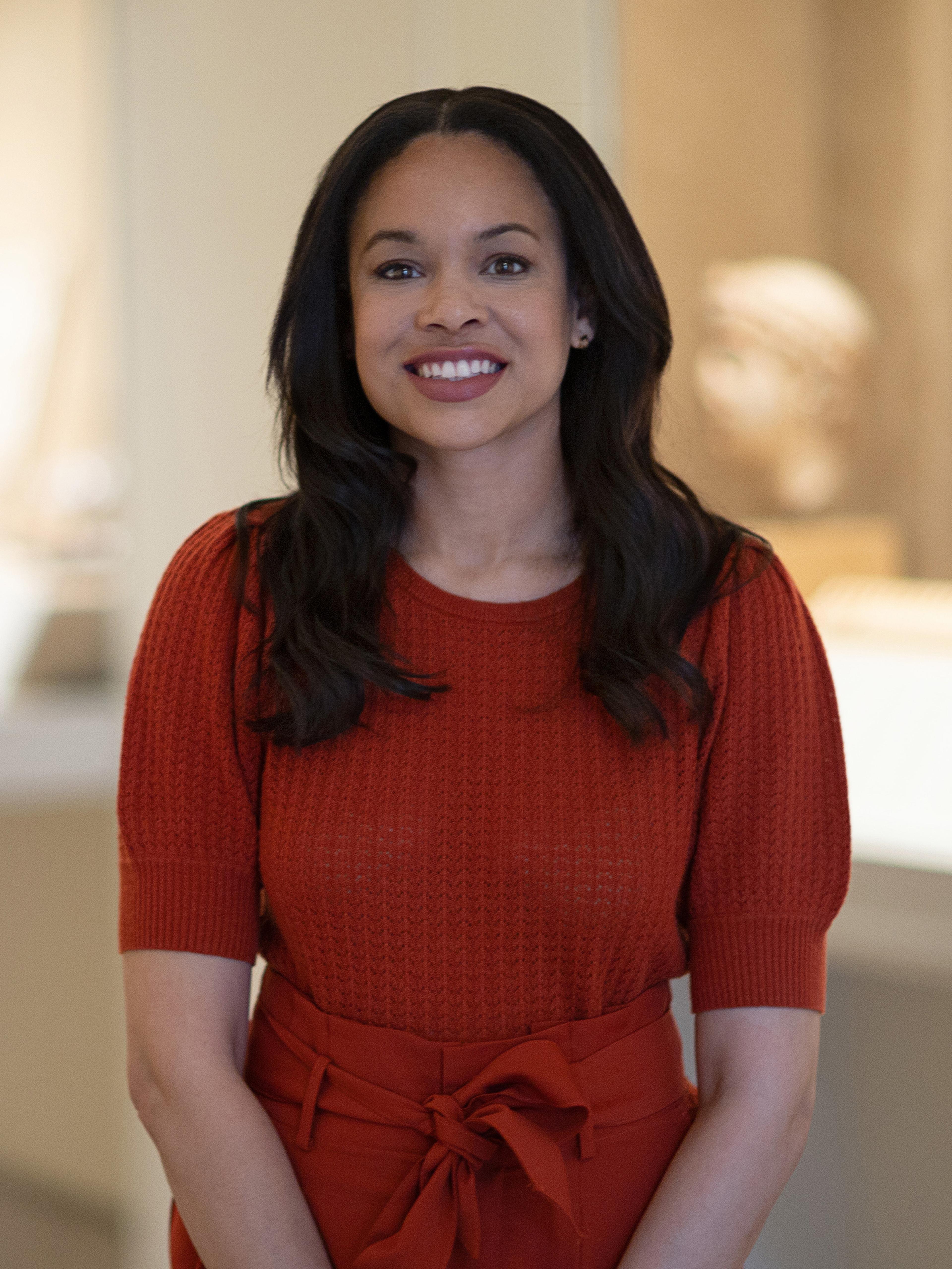A woman wearing a red blouse smiles in an art gallery