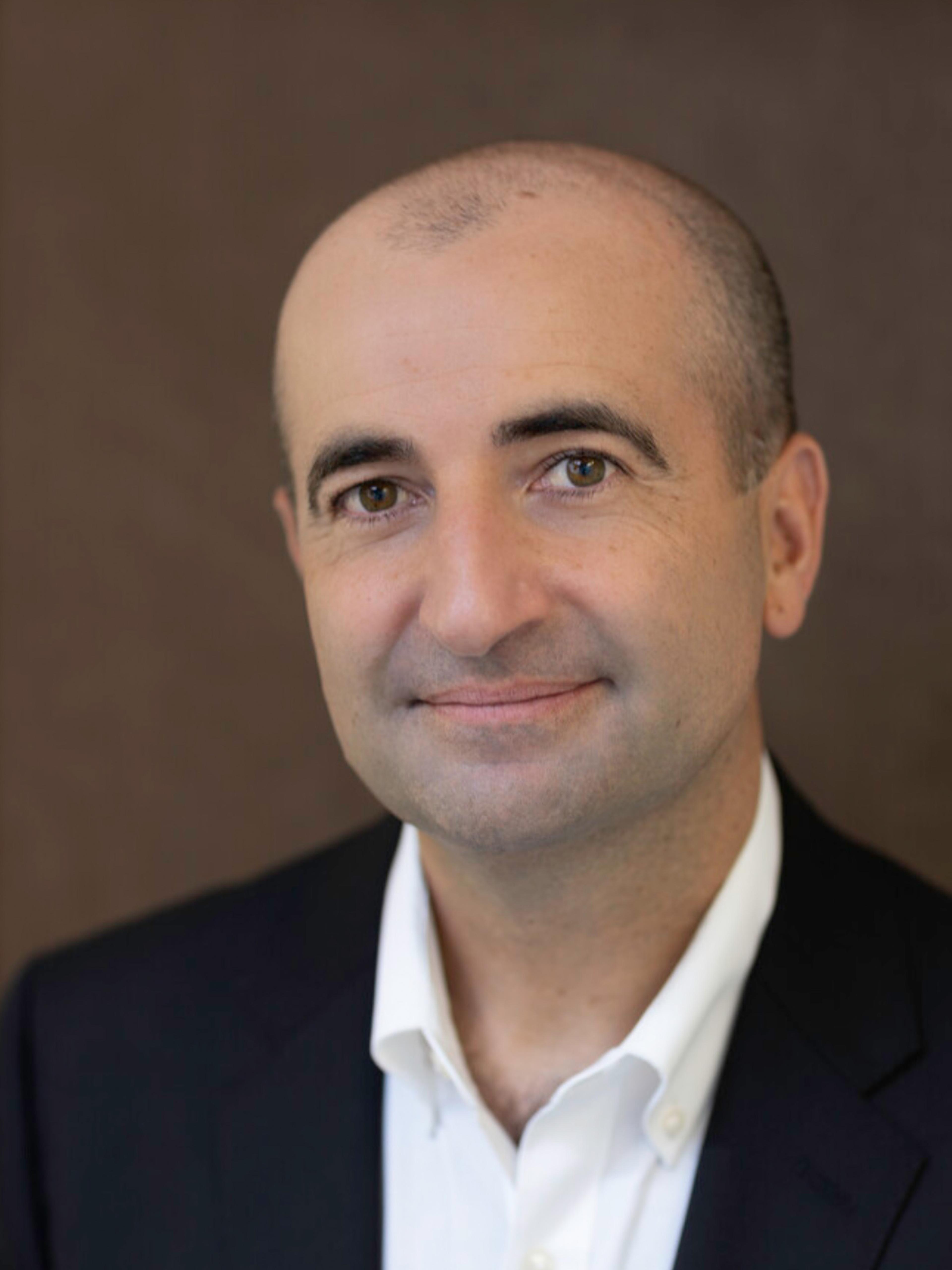 Professional headshot of a bald man in a dark suit and white shirt, looking at the camera with a calm, approachable expression against a brown background.