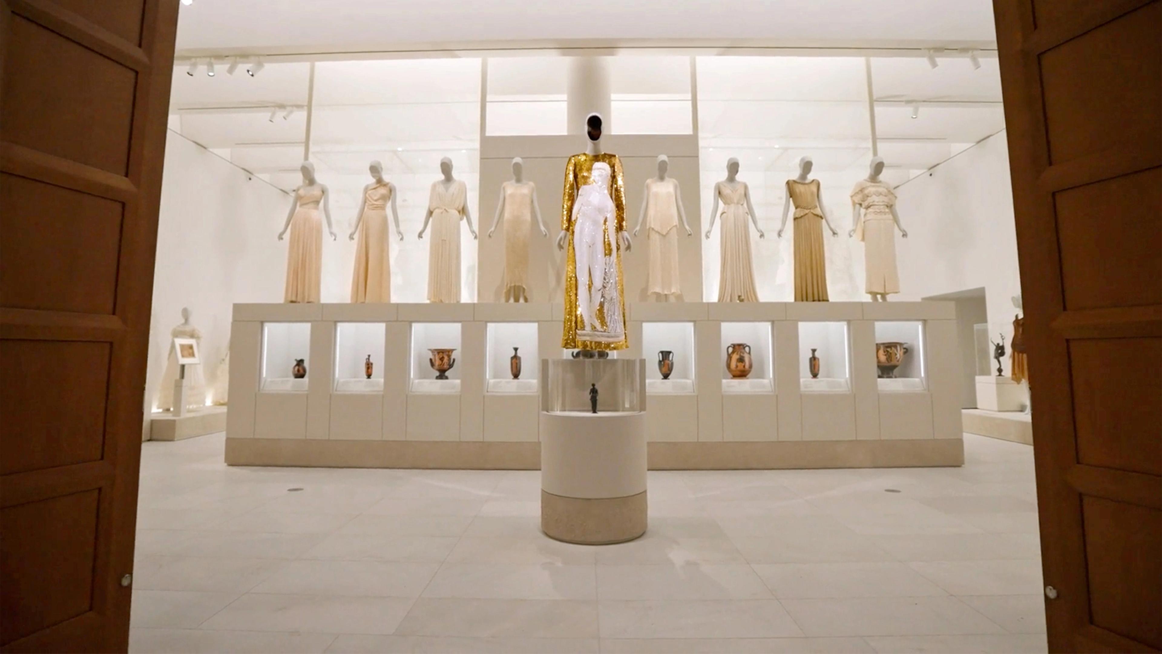 Tall wooden doors frame a high-ceilinged, bright gallery. A row of eight mannequins in beige gowns stand on vitrines in the background, while a central mannequin with a sequined gold dress stands on a pedestal in the foreground.