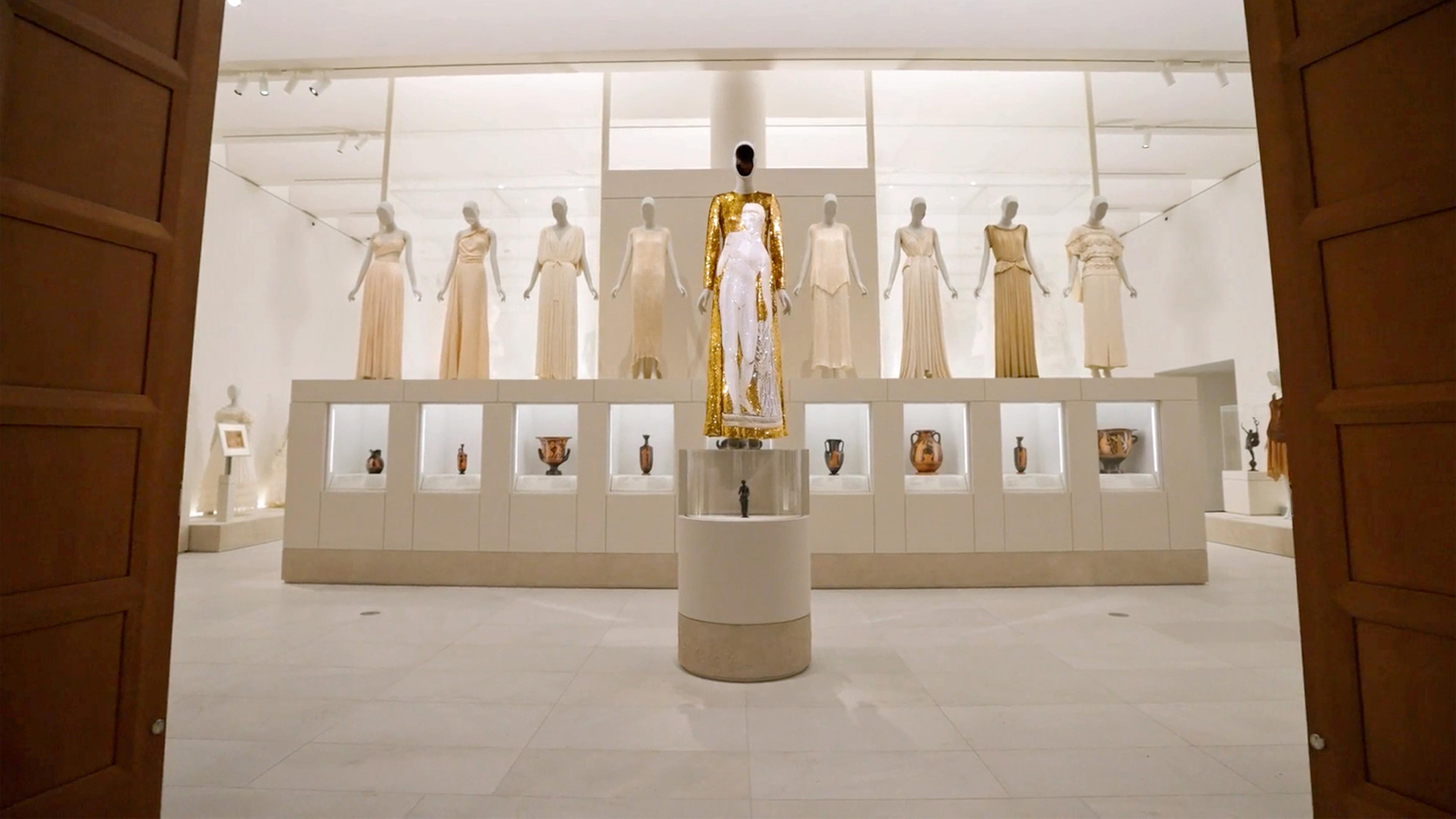 Tall wooden doors frame a high-ceilinged, bright gallery. A row of eight mannequins in beige gowns stand on vitrines in the background, while a central mannequin with a sequined gold dress stands on a pedestal in the foreground.