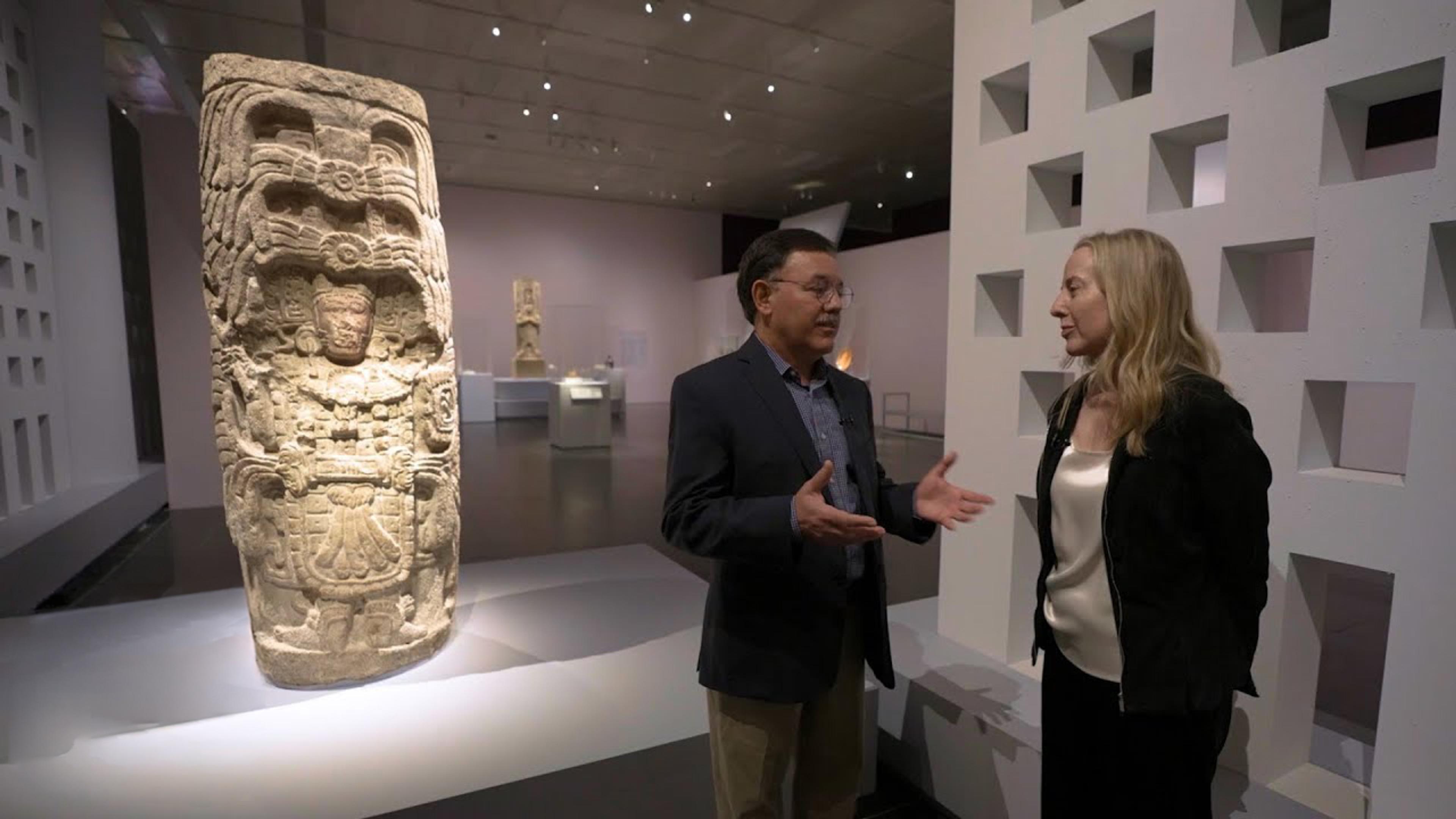 Two people stand in conversation beside a large carved Maya stone monument in a museum gallery.