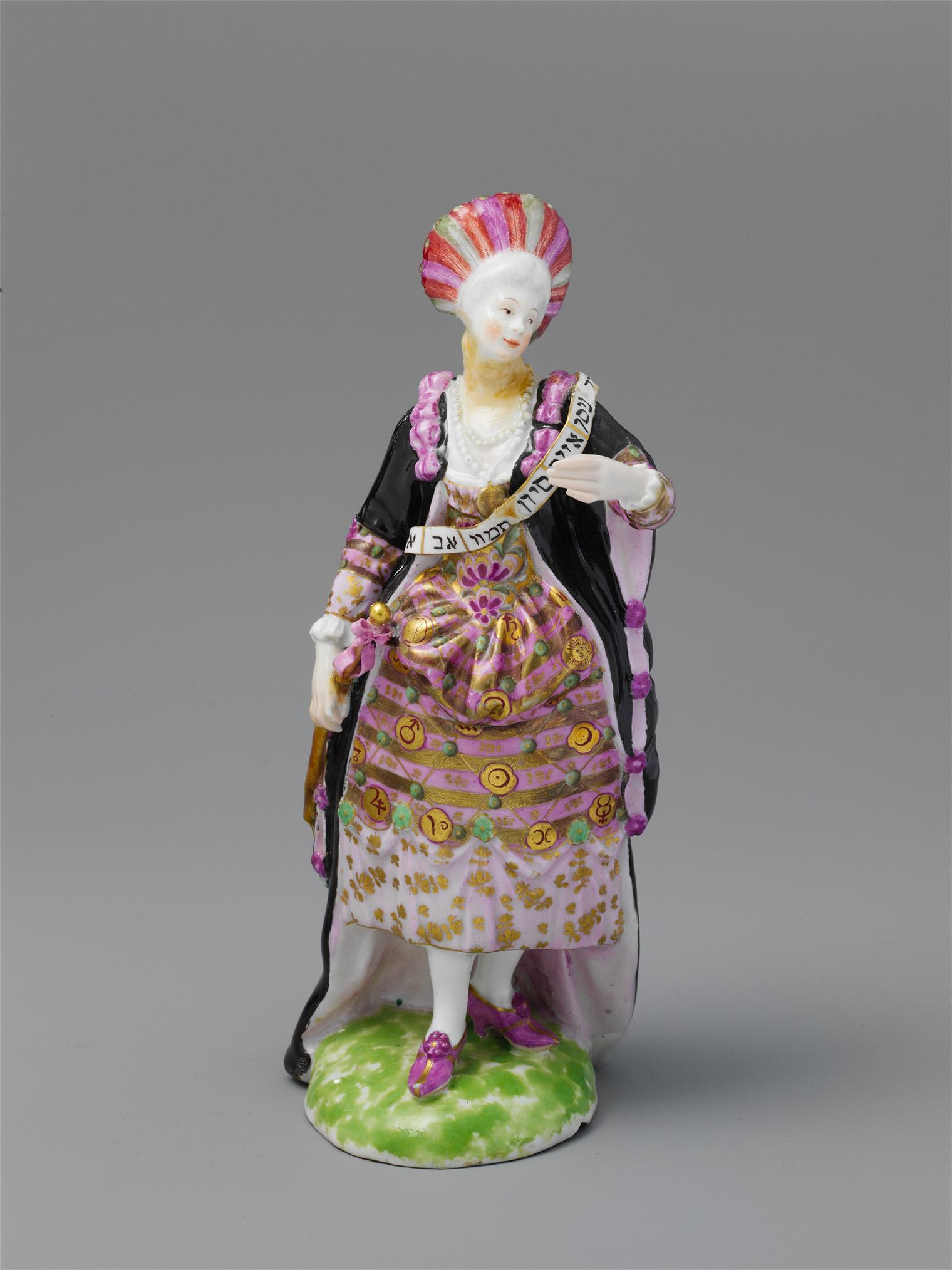 porcelain figurine of a woman in ornate 18th-century dress with pink and gold patterns, black cloak, and a fan. She stands on a green base.