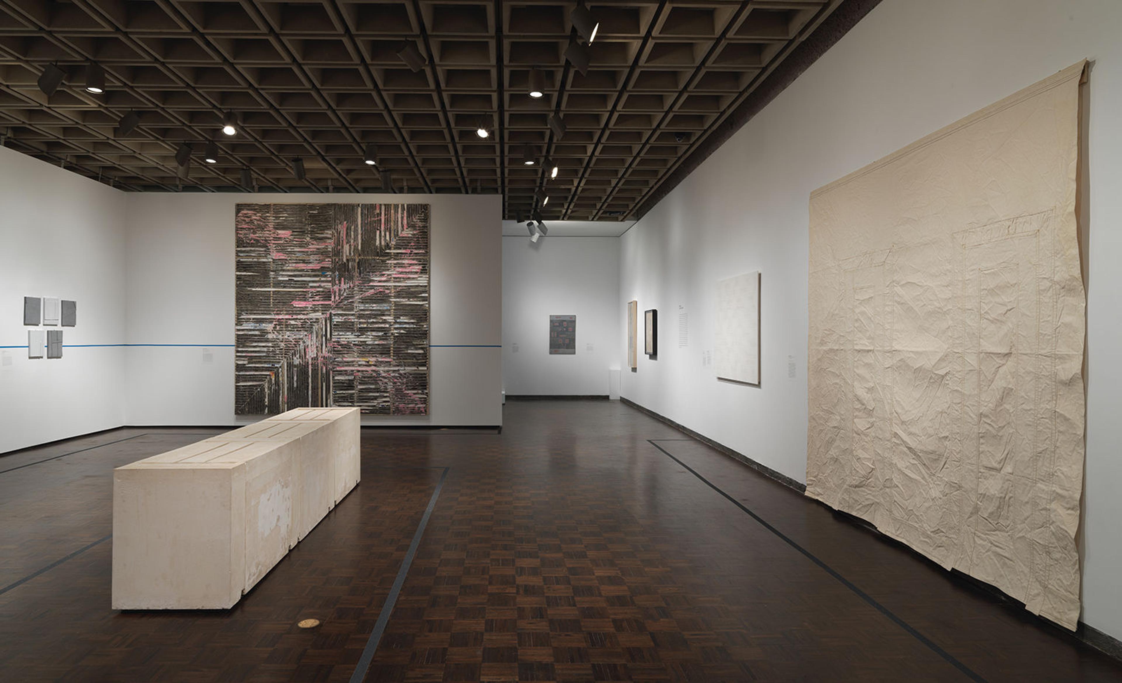 Gallery shot from Home is a Foreign Place. A bulky, cream-colored rectangular sculpture sits in the center of the checkerboard wood floor. Large-scale artworks hang on the surrounding walls.