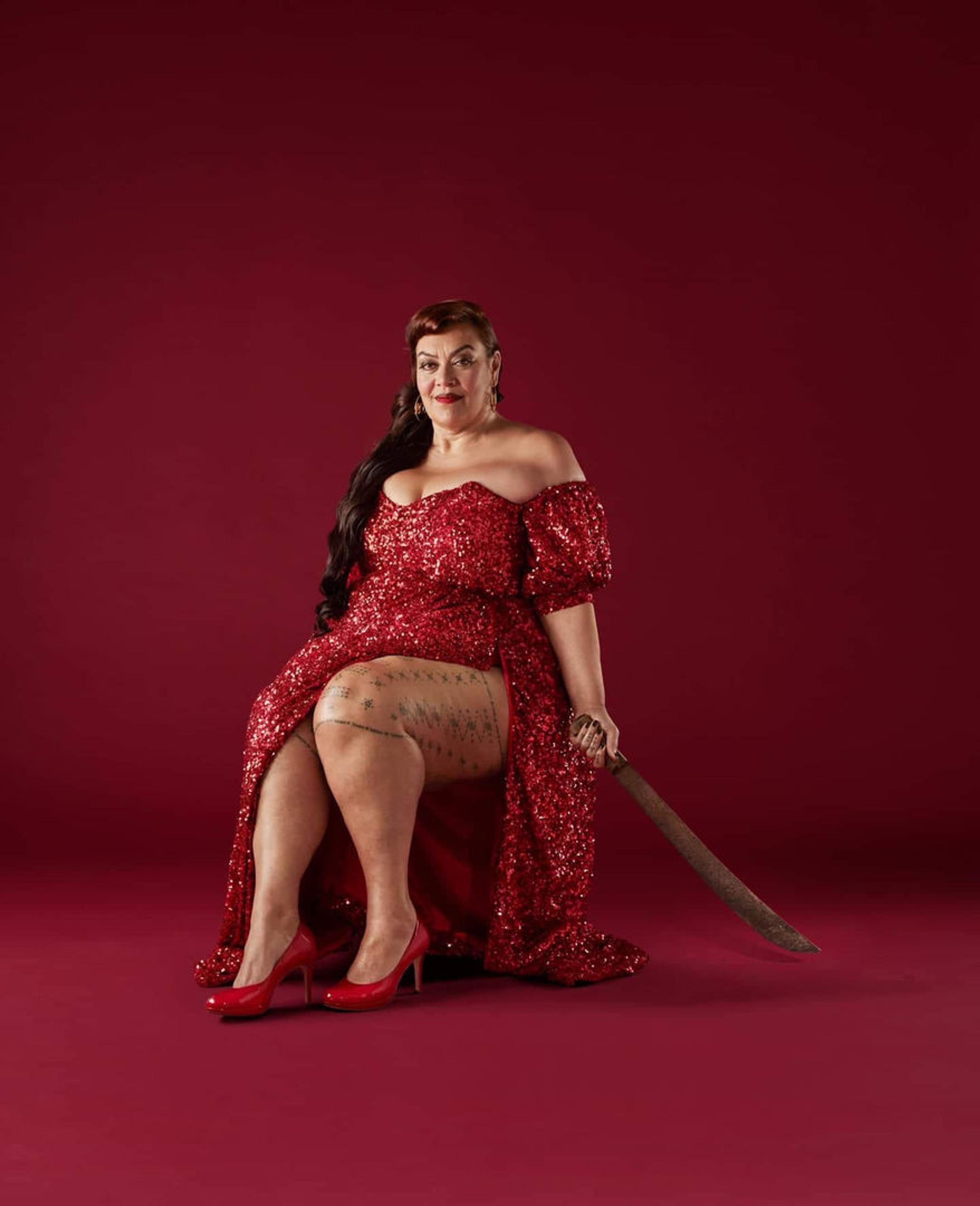 A woman with long dark hair wearing a red dress and red heels sitting against a red background holding a machete