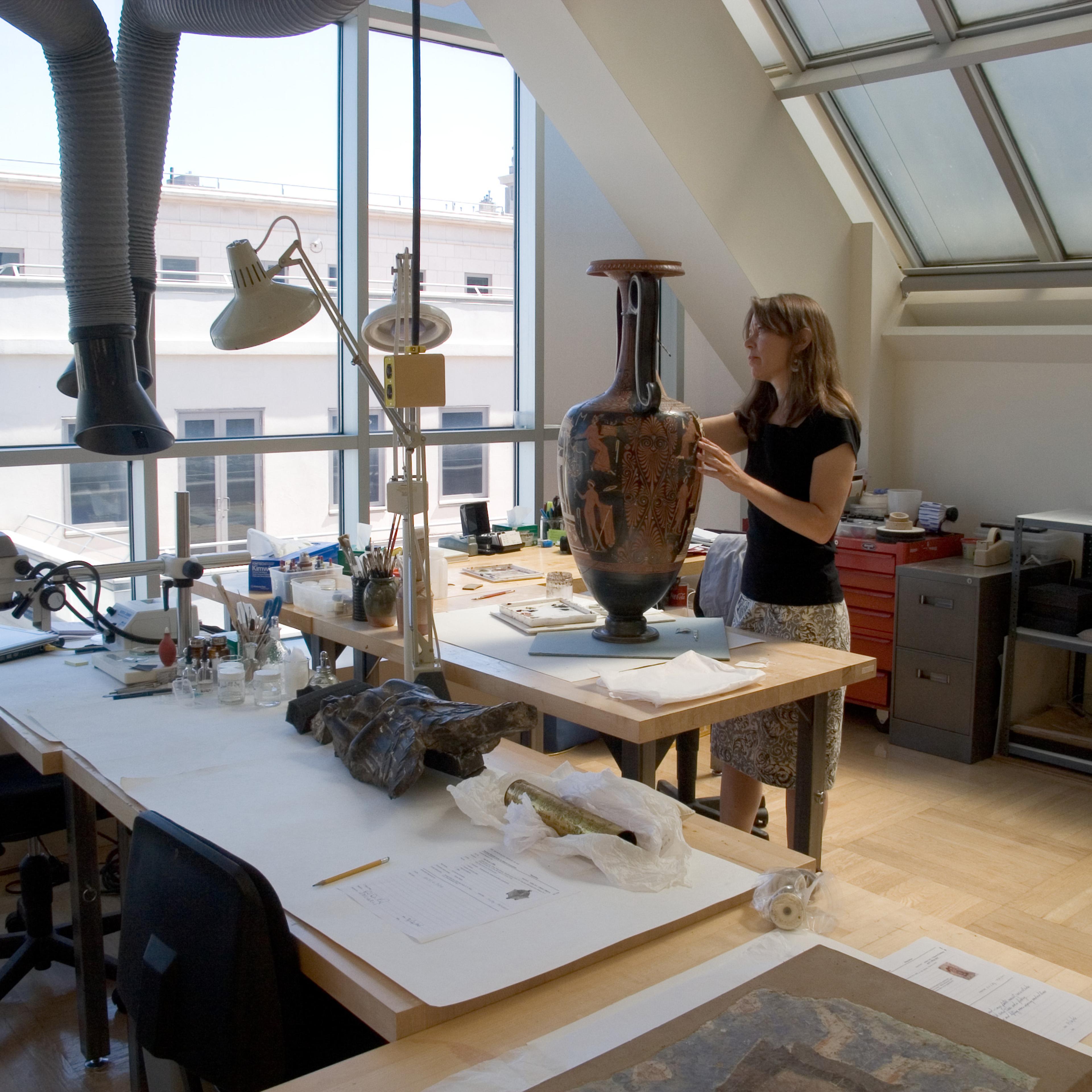 Conservator inside a brightly lit lab with large windows, works on a large vase