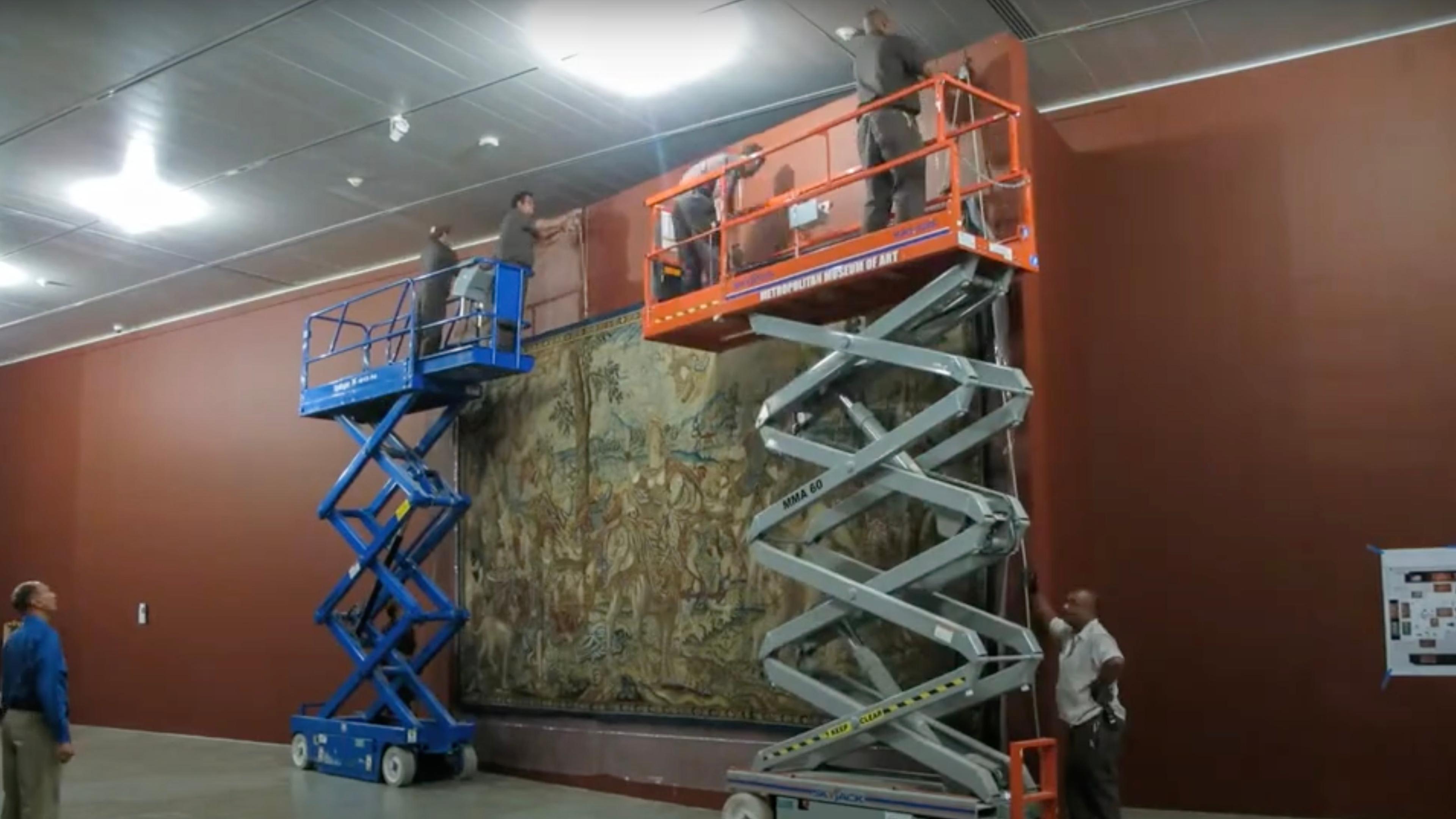 Four men elevated on two large scissor lifts installing a large tapestry in a gallery.