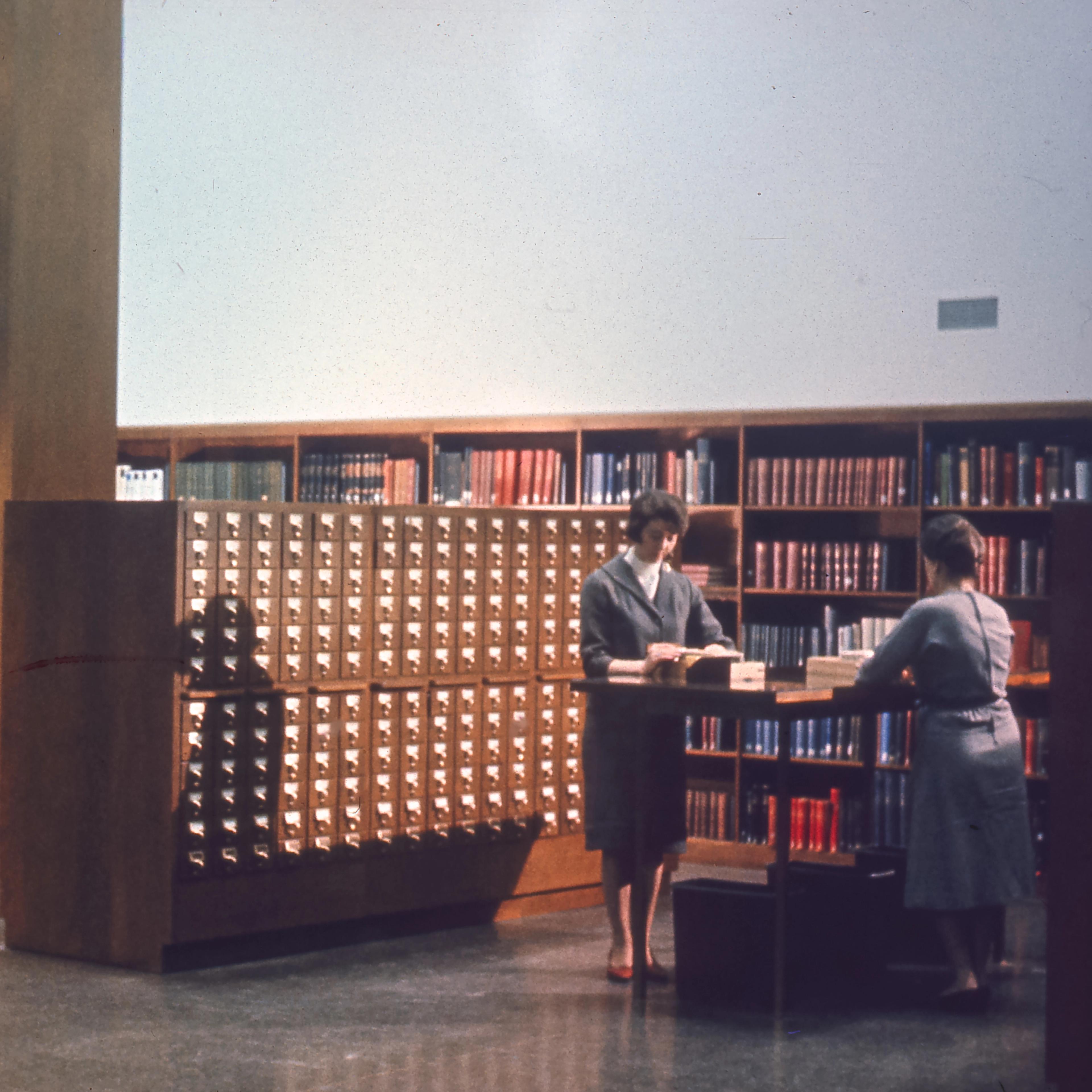 A spacious library with wood shelves filled with books. Two women are standing at a tall desk in front of a large card catalog working with books.