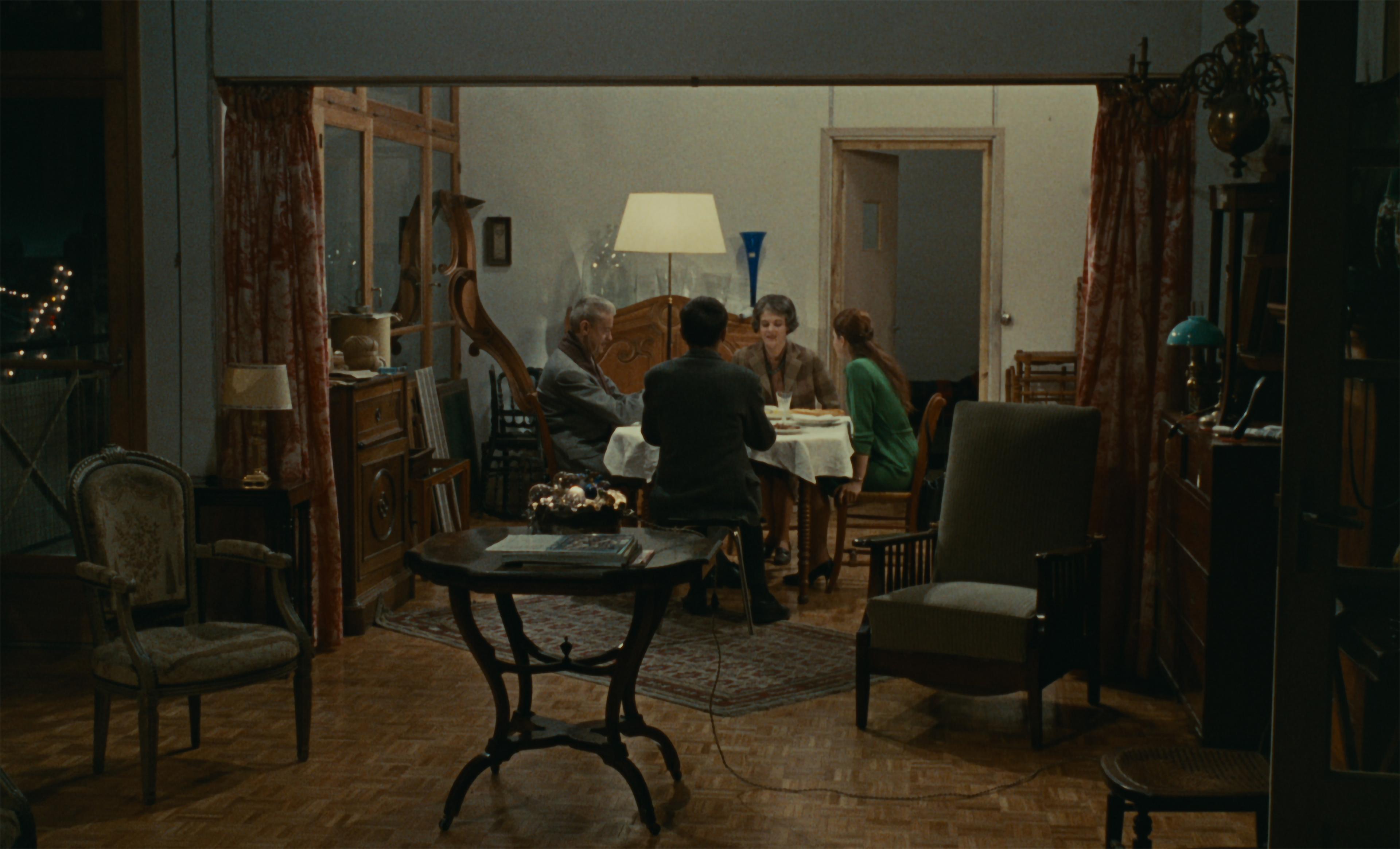 Scene from the French film Muriel. Four people sit around a table in a dining room framed by red and yellow curtains. In the foreground, a smaller table and two sitting chairs fill the parlor.