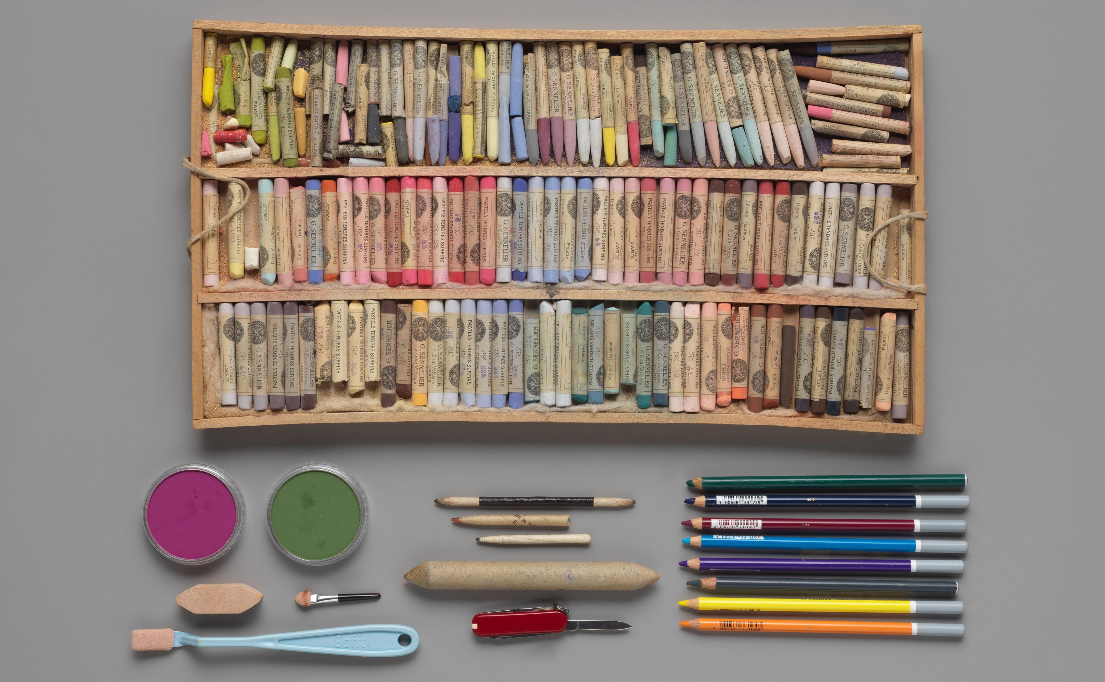 A wooden box of dozens of colorful pastel pencils, above an arrangement of other drawing implements.