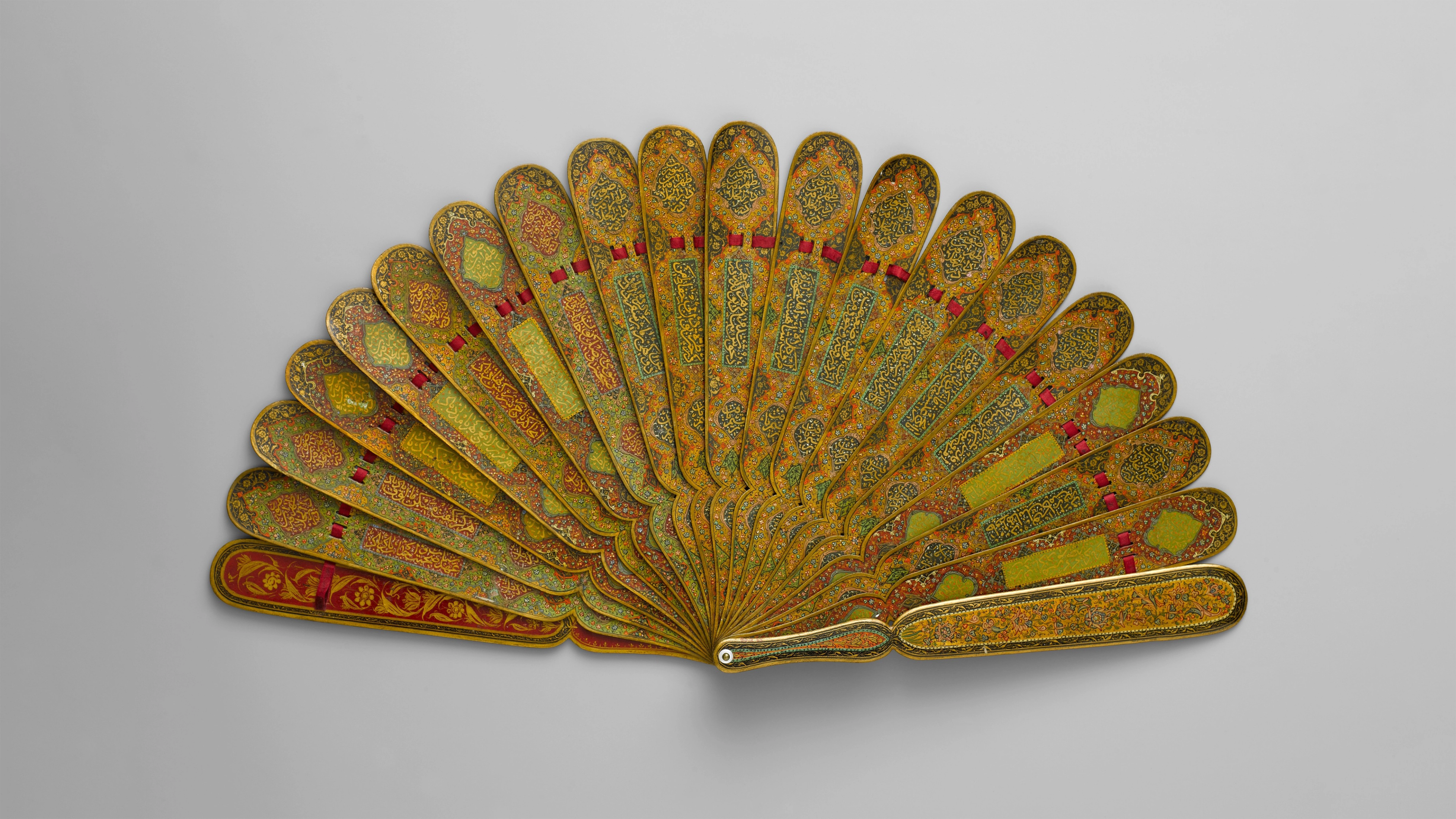 Ornate folding fan with golden patterns and red accents on each rib, intricately designed with floral motifs, displayed against a neutral background.